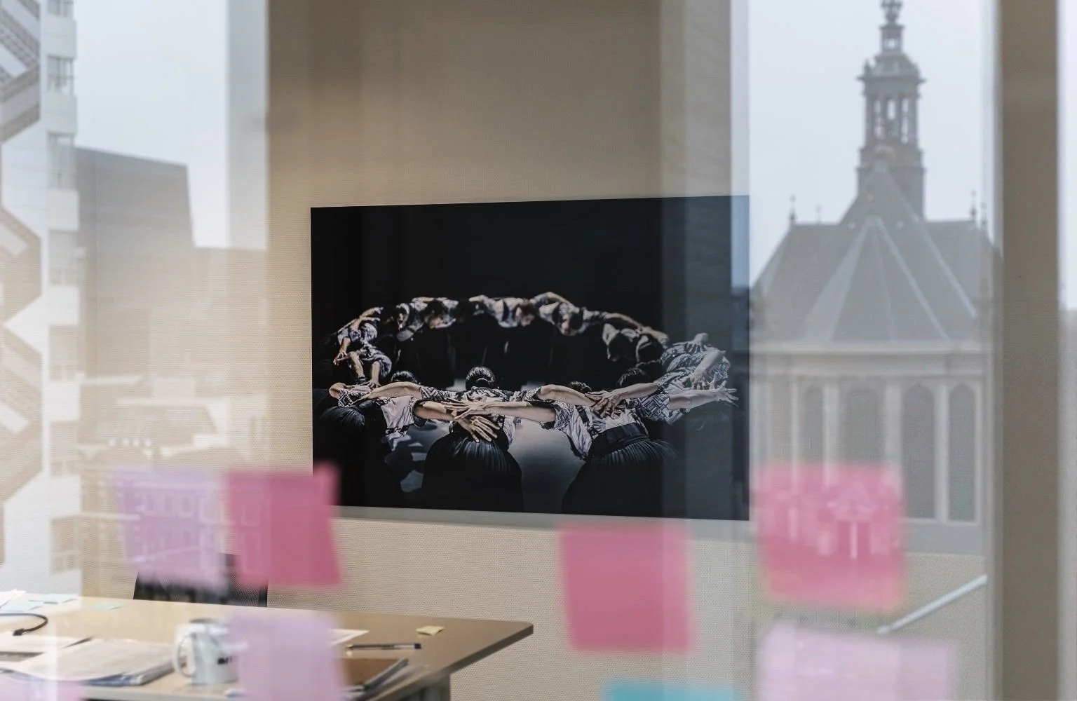 Een photo van een kunstwerk met mensen die een cirkel vormen, die op een muur wordt tentoongesteld, gezien door een raam met roze post-its and een grote raam van een gebouw met een toren erachter.