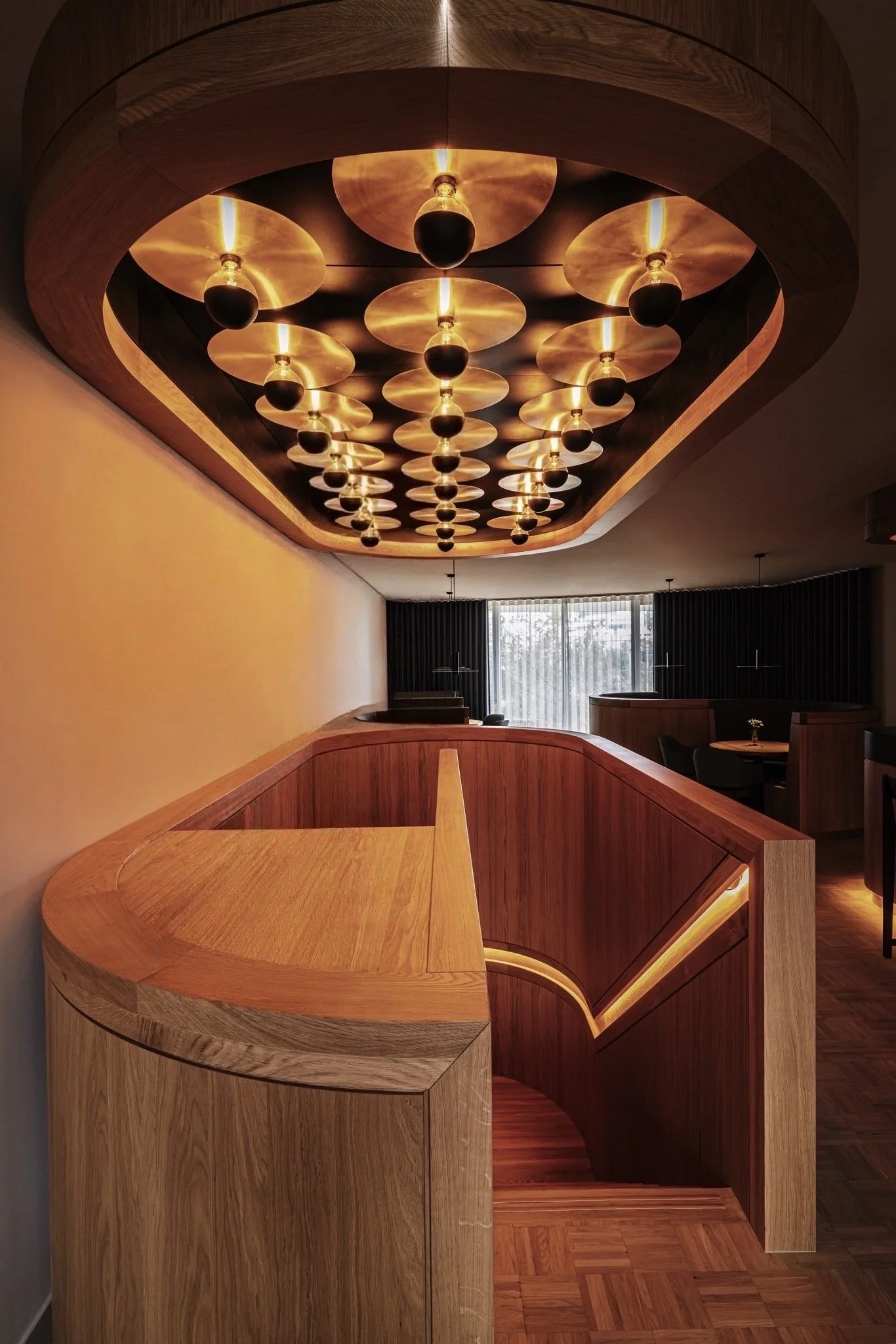 Een houten balie met een ronde rand en een opengewerkte zijkant, onder een houten plafond met een moderne lichtinstallatie bestaande uit meerdere hangende lampen met ronde vierkante schijven.