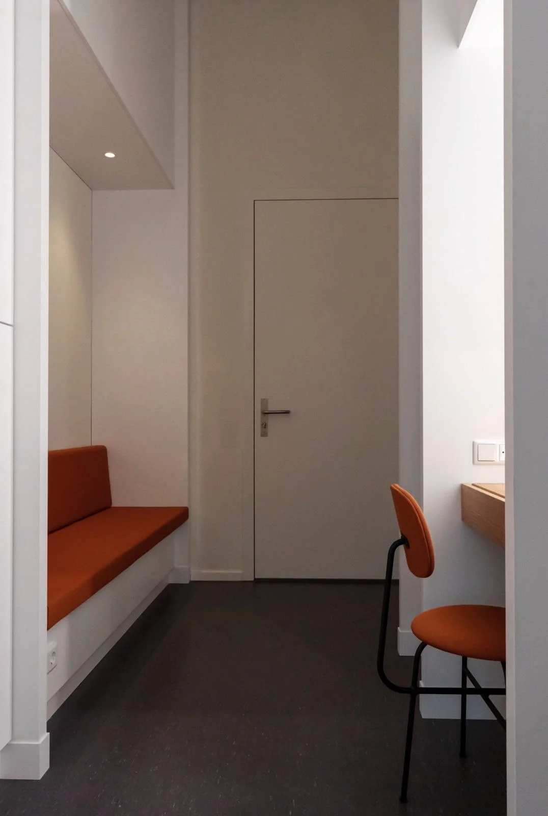 Een rustige, moderne kamer met een deur, een bankje met oranje bekleding aan de muur, een stoel met oranje bekleding en een klein houten bureau in een lichte, minimalistische inrichting.