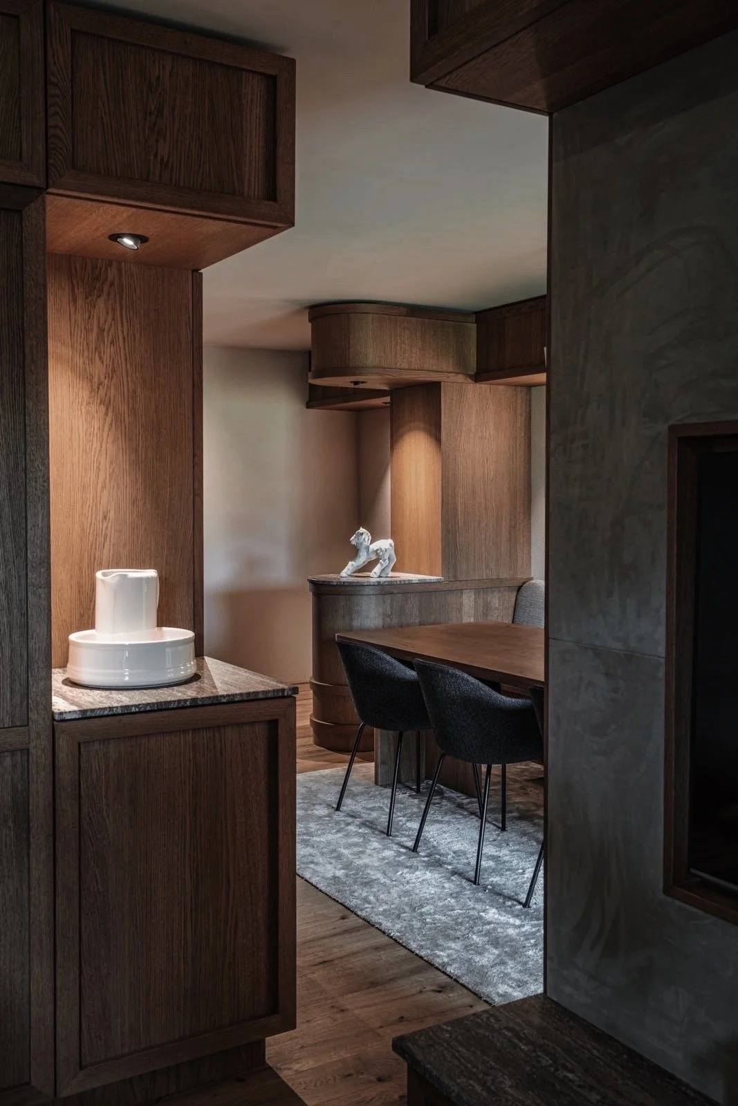 Binnenkant van een huis met houten meubels, een zwart tafelblad met drie zwarte stoelen, een tapijtvloer en decoratieve objecten zoals een witte pot en een sculptuur van een hond.