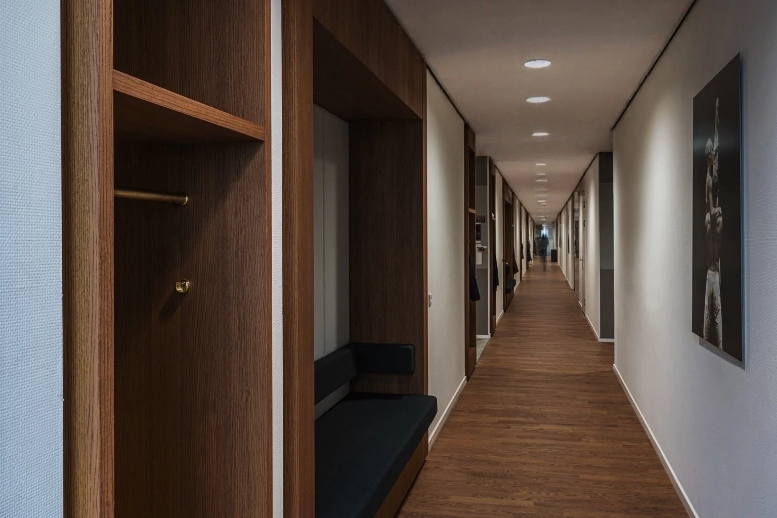 Lange, smalle gang in een moderne binnenruimte, met houten vloer en lichte muren. Aan de linkerzijde zijn open kasten en een zwart bankje, aan de rechterzijde hangen foto's. Verlichte plafondspots leiden naar de diepte van de gang.