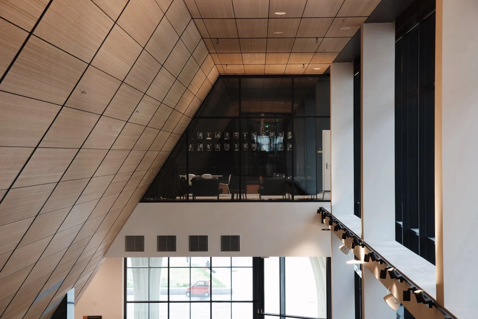 Interieur van een modern gebouw met een houten plafond en glazen wanden, met een ruimte boven met zwart-witte portretten aan de muur.