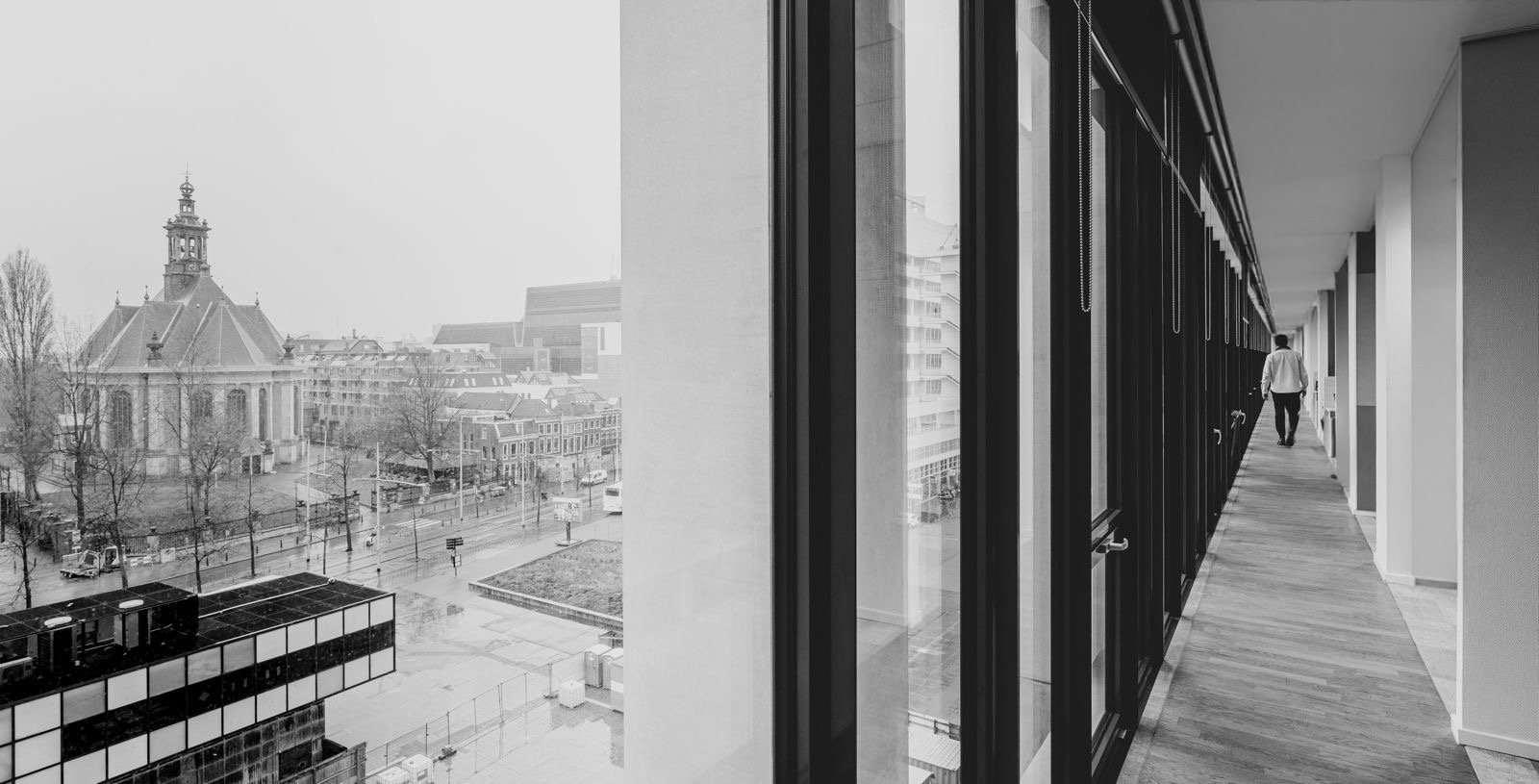 Binnenzicht van een gang bij de kantoren van Nederlands Dans Theater in Amare aan Spuiplein in Den Haag met een lange gang en ramen die uitkijken op een stad met historische en moderne gebouwen.