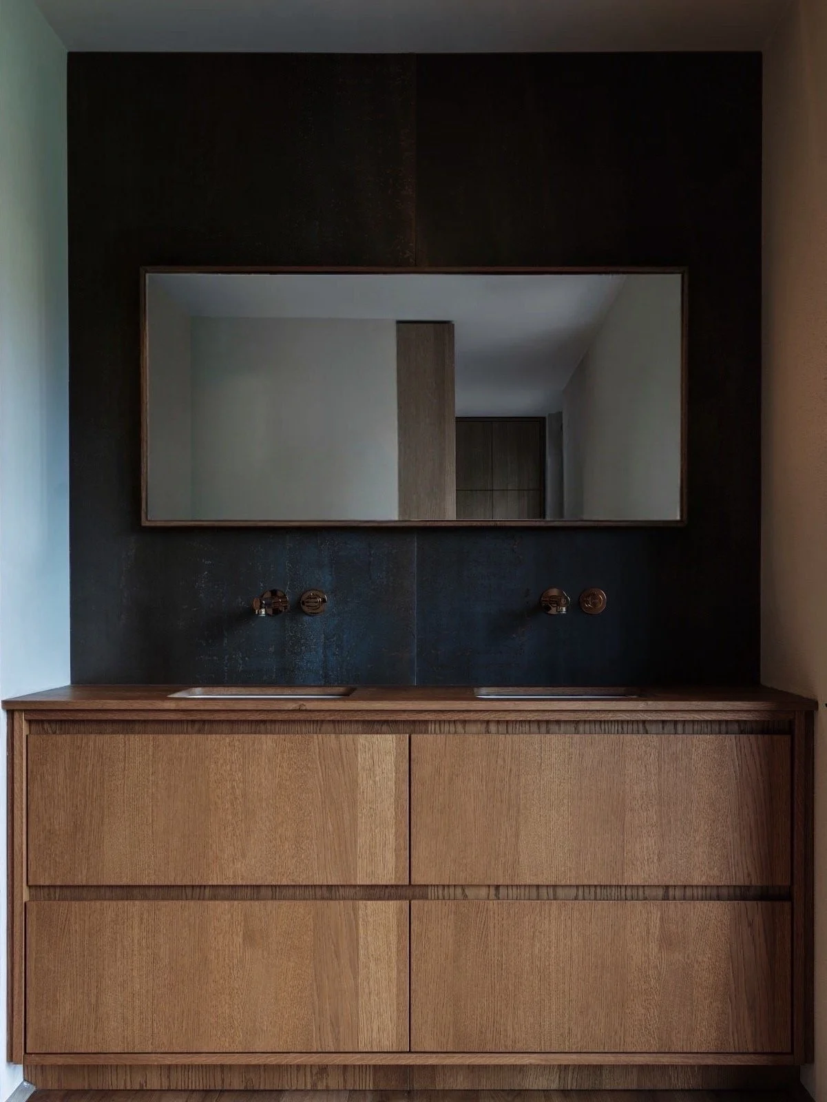 Een houten badkamermeubel met twee wastafels en een groot spiegel erboven, tegen een donkere muur.