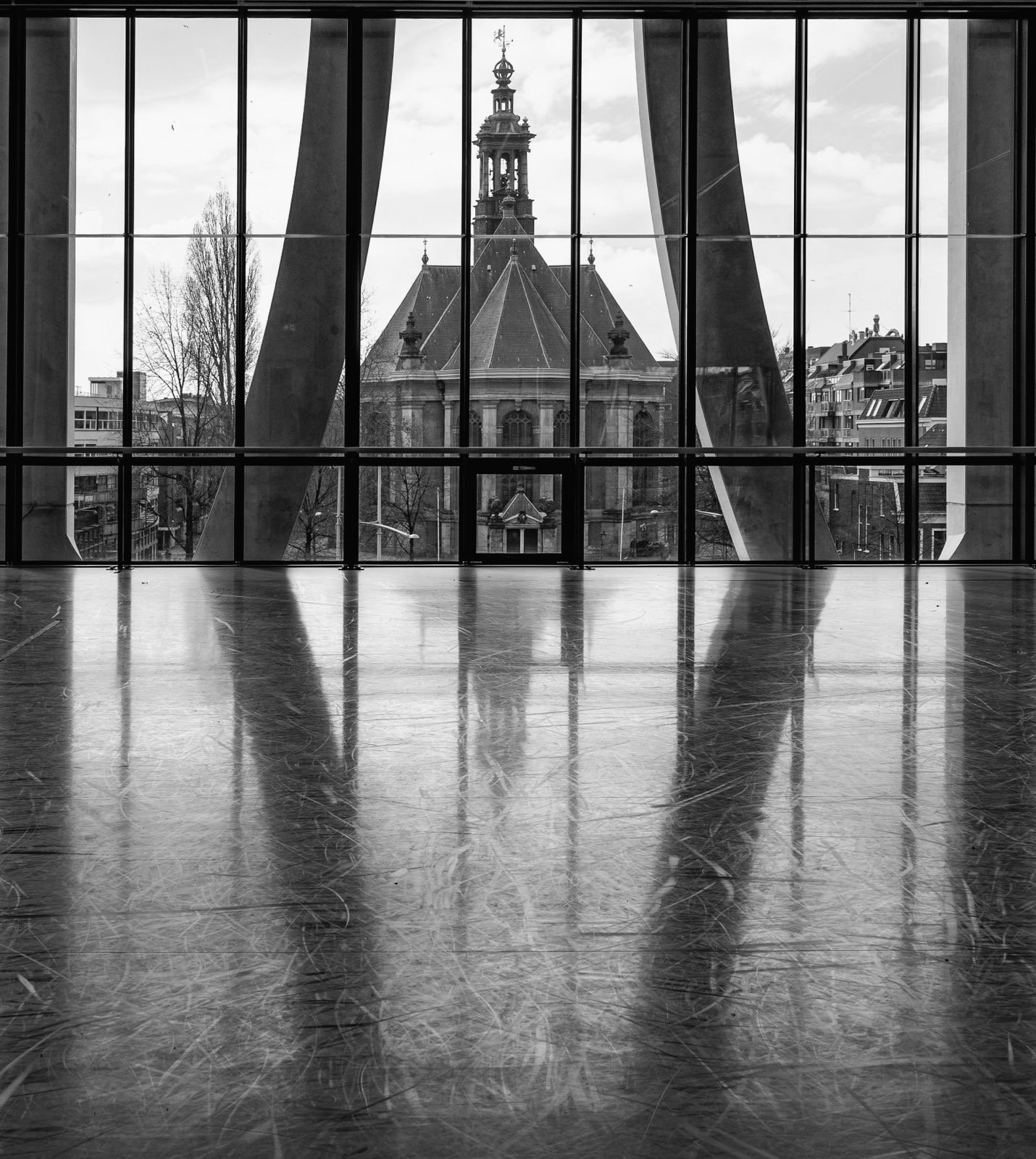 Binnenzicht van een moderne, lege, glanzende houten vloer met grote ramen die uitkijken op een historische kerk of gebouwen met een toren, onder een bewolkte lucht.