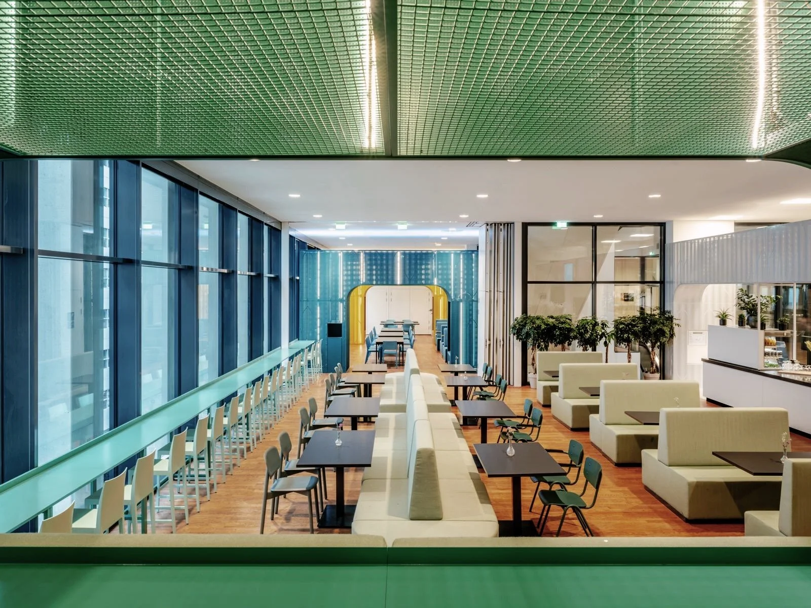Interieur van een modern restaurant of café met tafels, stoelen, groene planten en grote ramen met uitzicht naar buiten.