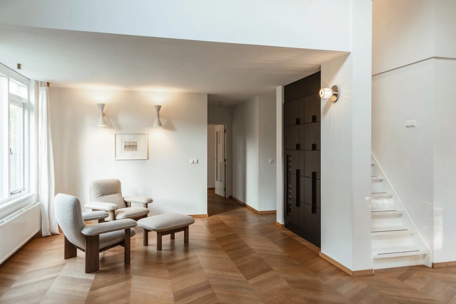 Woonkamer met eiken houten vloer in Hongaarse punt, twee beige Audo Brasilia stoelen en een zwarte maatwerk kast element, witte muur, raam met wit gordijn, design muurverlichting van Le Corbusier en DCW Editions, en een witte trap.