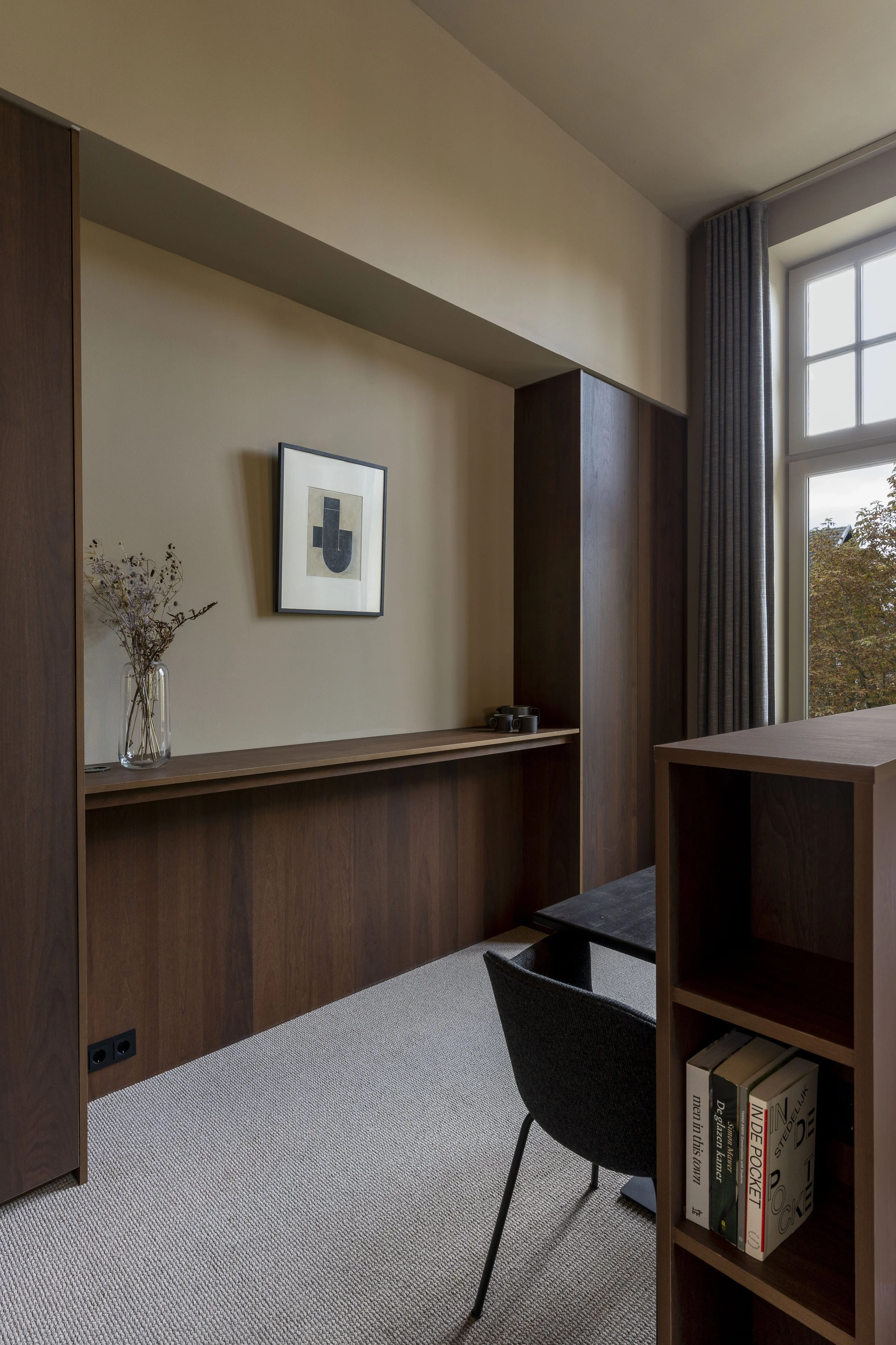 Een interieur van een rustige kamer met een grote ramen, een houten bureau, een zwarte stoel, een boekenkast met boeken, een kunstwerk aan de muur en een vaas met bloemen.