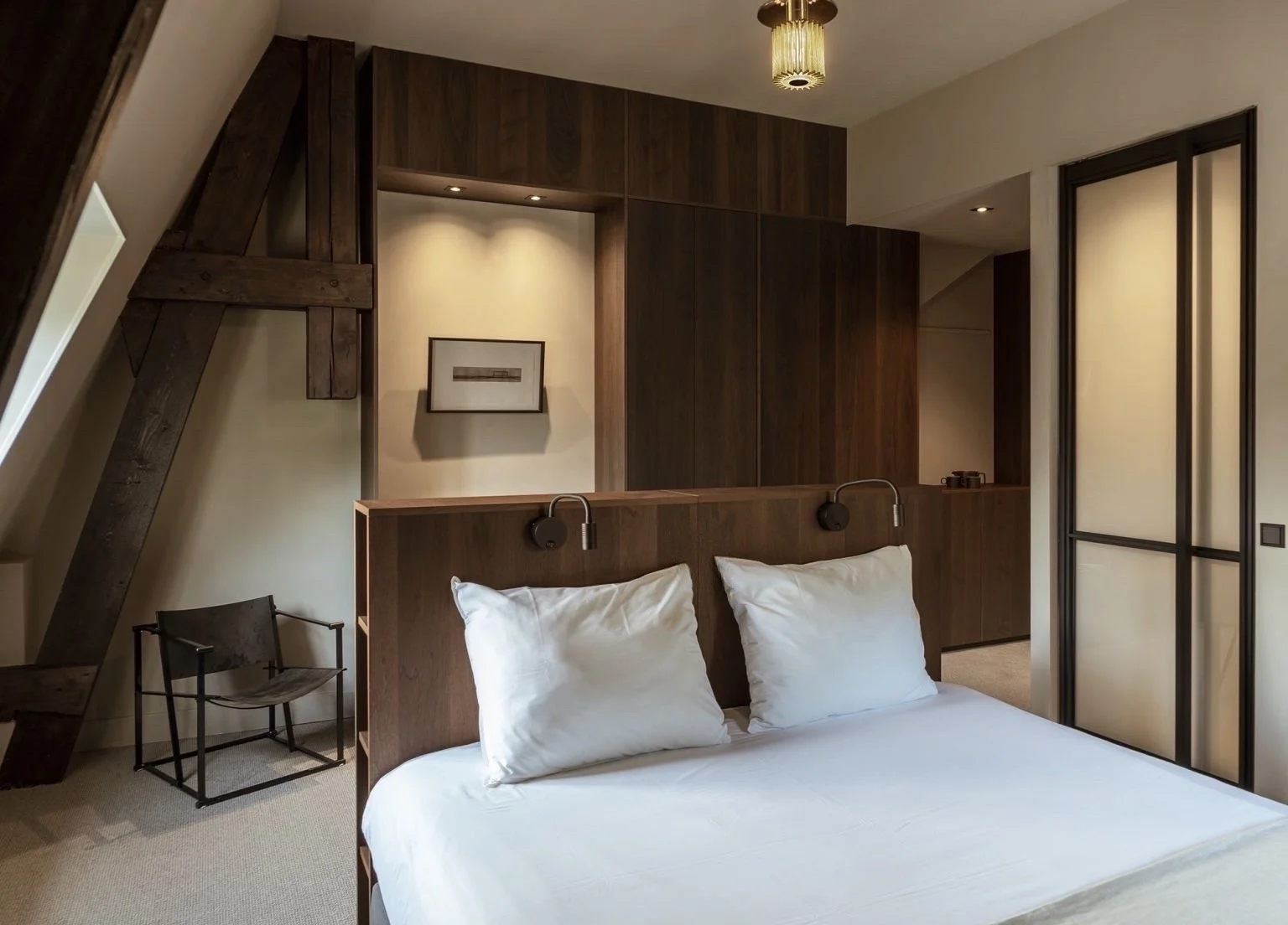 Slaapkamer met wit bed, houten scheidingswand, schouderlampen, zwart metalen stoel, en een decoratief plafondlamp.