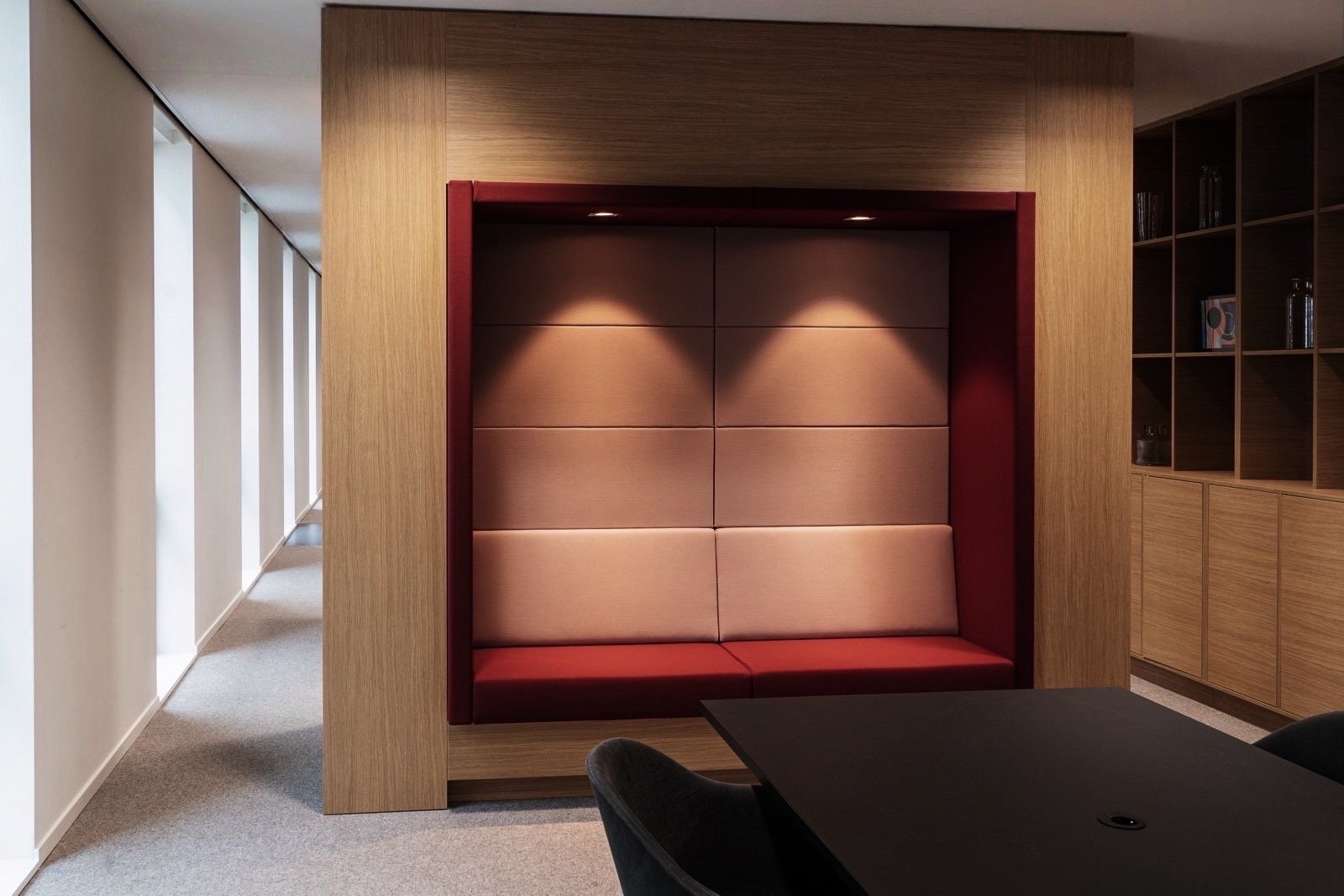 Binnenkant van een modern kantoor of hotel met een comfortabele zitruimte met hoge, gewatteerde bekleding in beige en rood, ingebouwd in een houten structuur. Aan de rechterkant een houten boekenkast en een tafel met een zwarte stoel.