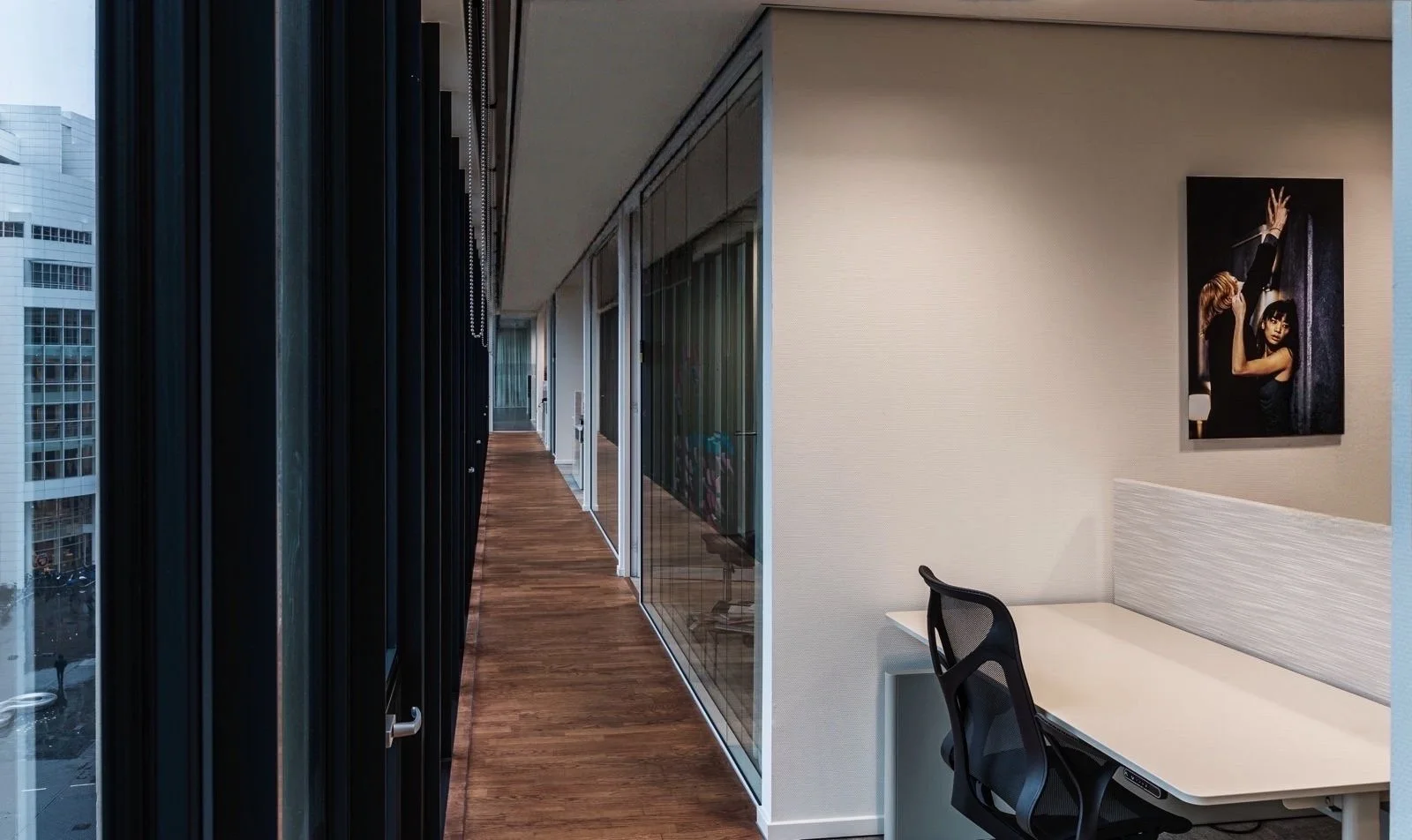 Kenniskantoor met corridor, glazen wanden, kunstwerk met twee vrouwen op de muur, en werkplek met bureau en stoel.