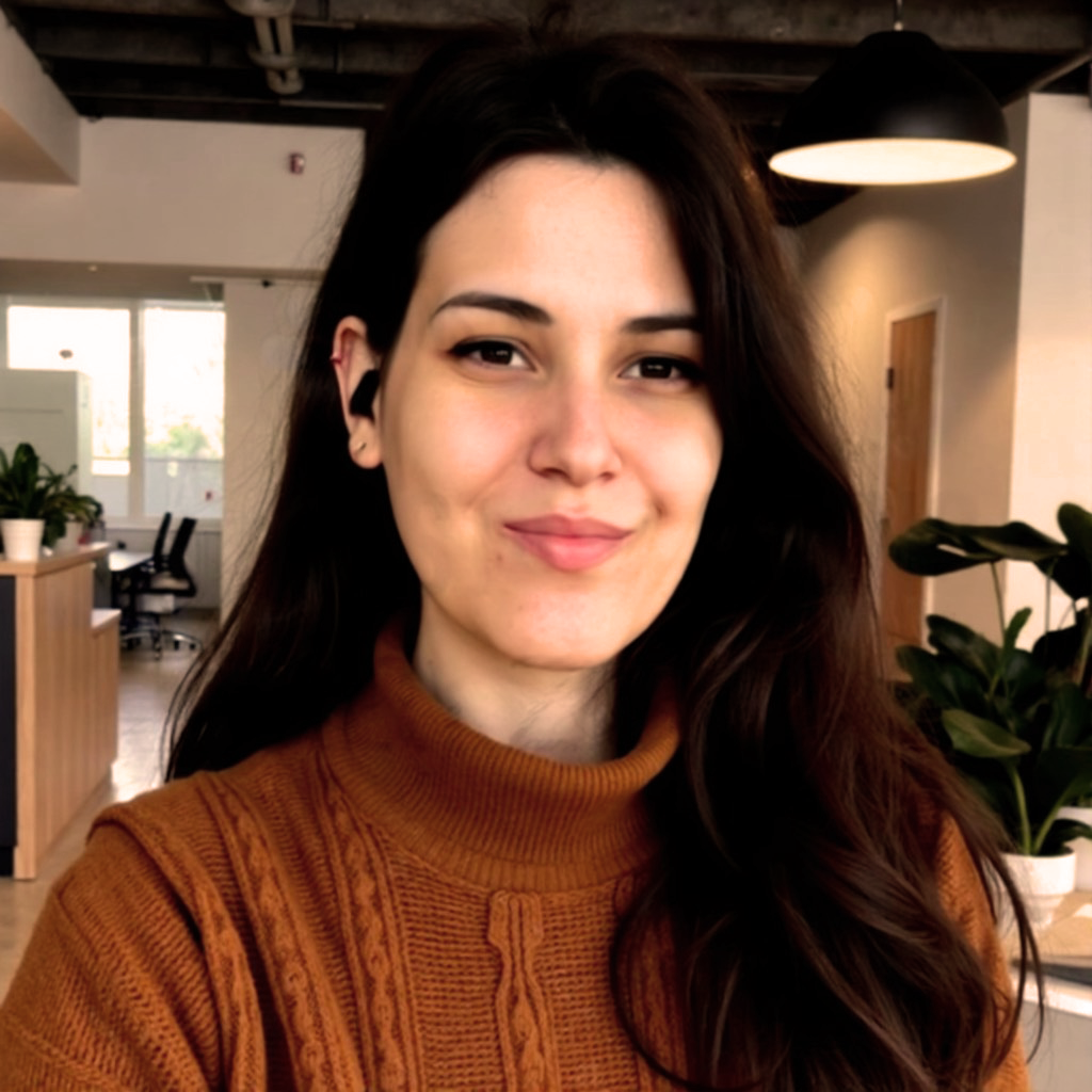 Portrait of a young woman with long dark hair, wearing a brown sweater, smiling indoors with modern decor and plants in the background.