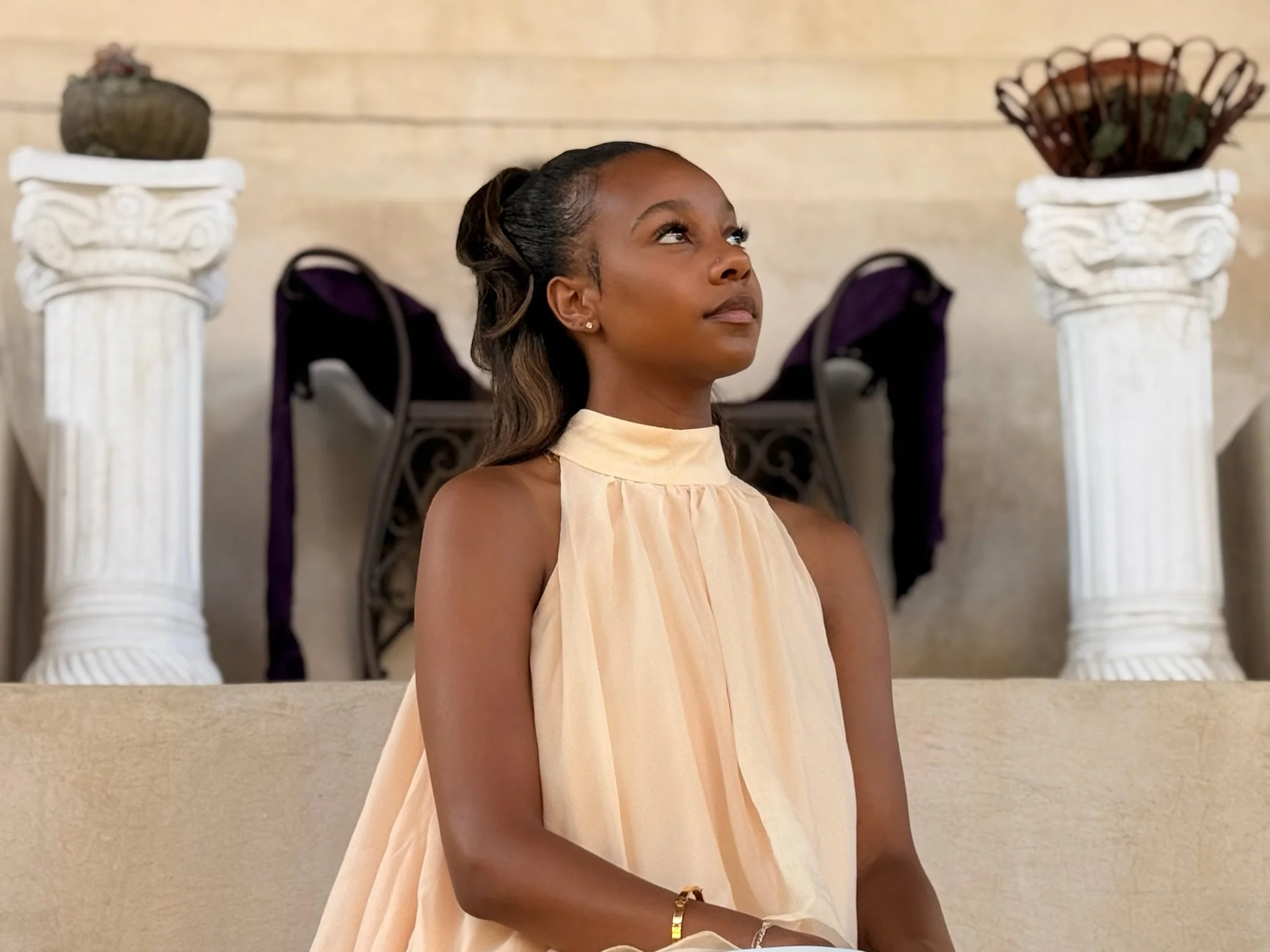 A young woman with dark skin and wavy hair styled to one side, wearing a light beige dress with cutout shoulder details, sitting on a beige staircase between two white classical columns with decorative capitals. There are two planters on top of the columns, one with brown and green plants and the other with dried plants, against a cream-colored wall.