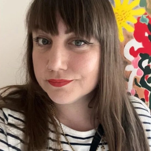 A woman with brown hair and bangs, wearing red lipstick, a nose ring, and a striped shirt, standing in front of a colorful floral background.