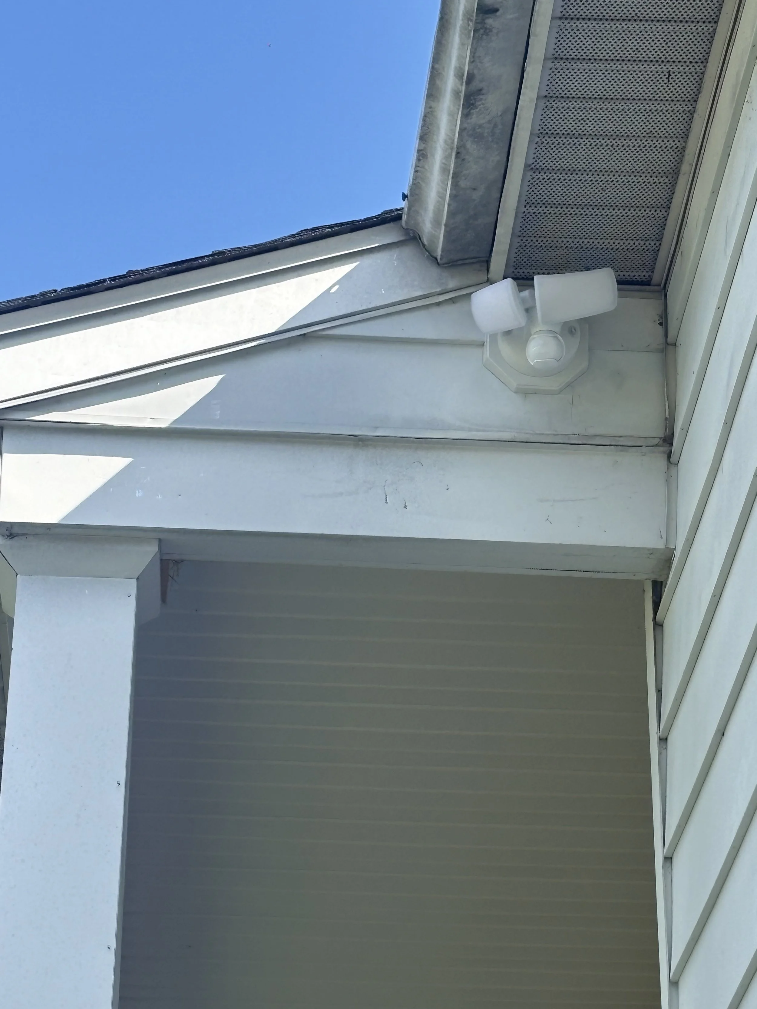 Close-up of a white outdoor light fixture mounted on a wall beneath a window with a window screen, with a clear blue sky in the background.