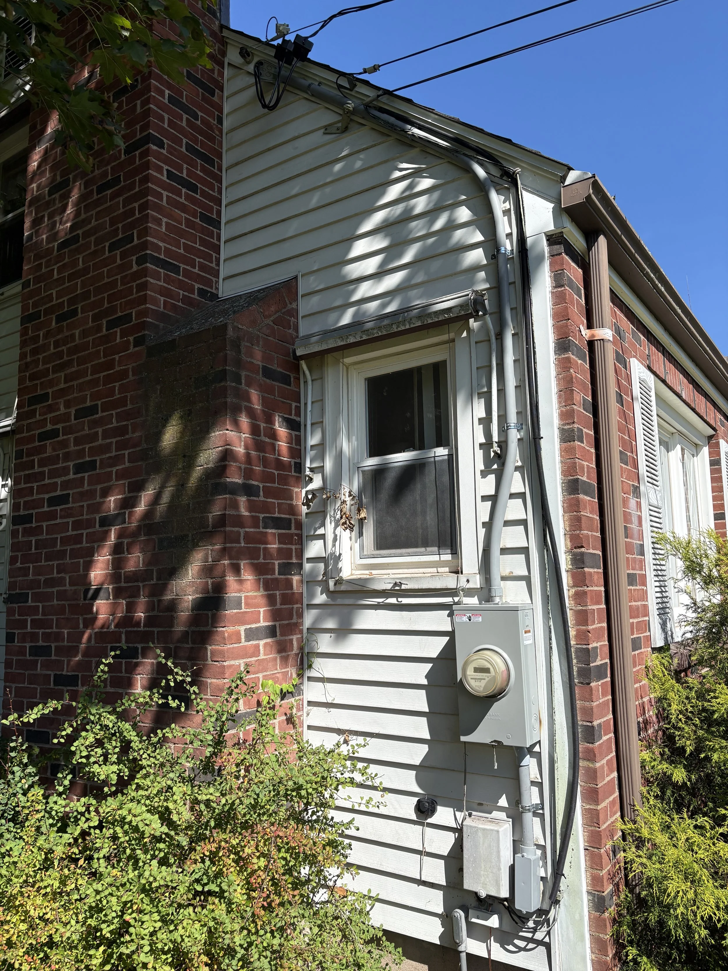 The image shows the exterior corner of a house with a brick wall and white vinyl siding. There are visible electrical and cable wires on the house's upper side, along with a gray electrical meter box and various conduits attached to the wall. Part of a window with a screen and white shutters is also visible. The scene is under bright sunlight, with shadows cast by nearby foliage.
