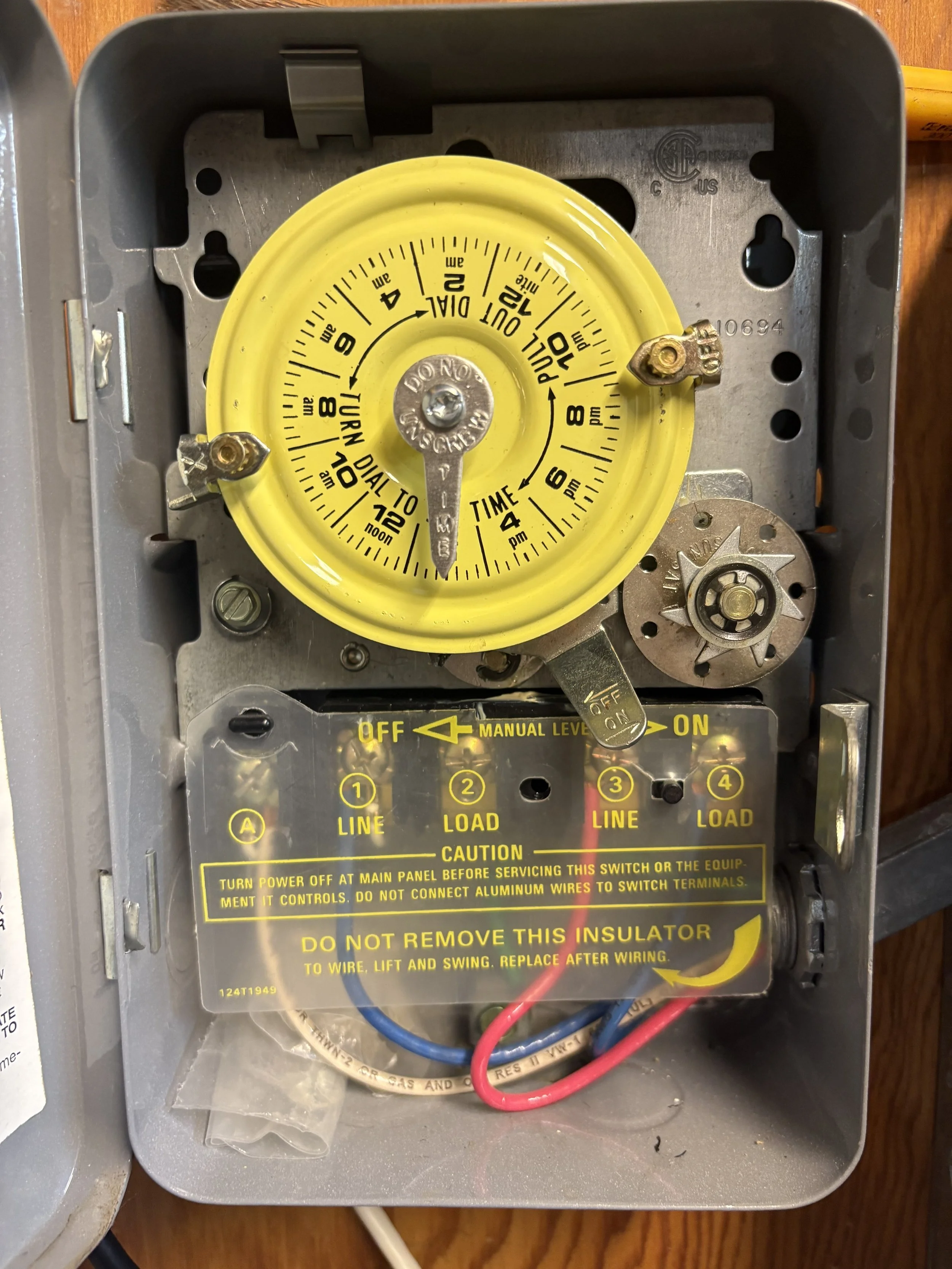 An electrical control box with a yellow dial for setting a timer, labeled in 24-hour and 12-hour formats, with instructions and warning labels below. The box contains wires and switches for controlling a device.