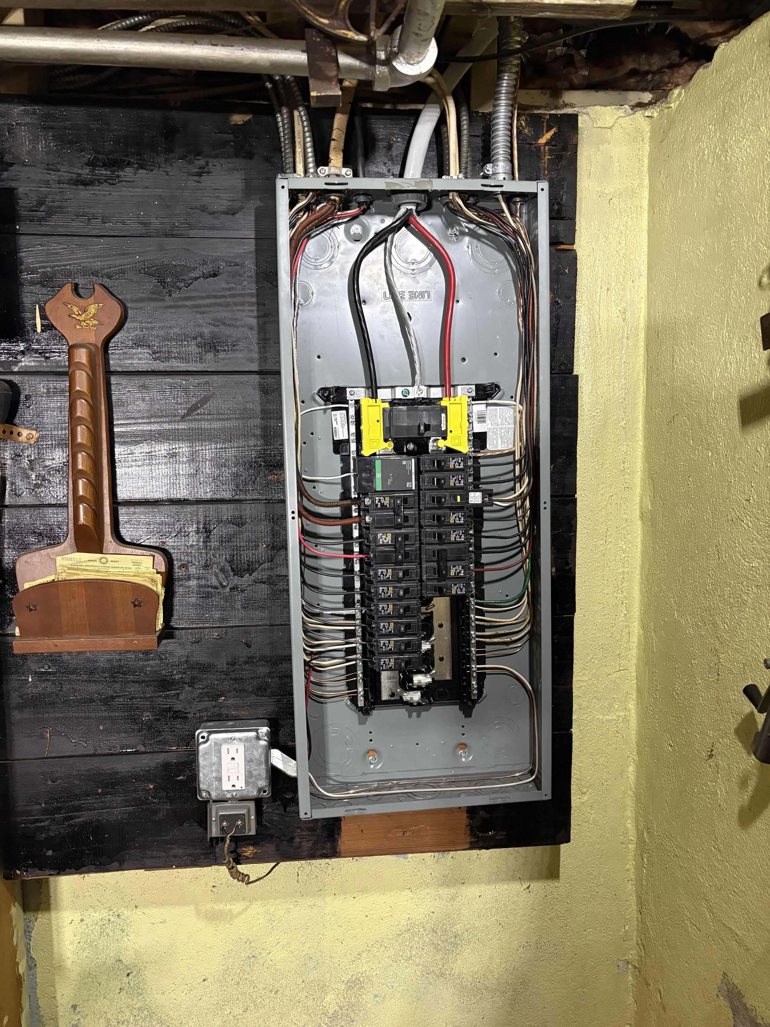 Open electrical panel with wiring and circuit breakers, mounted on a wall with black and yellow paint. A large wrench is on the left side and a power outlet with plug below. The wires installed in the panel are neat and orderly. CT NEC Compliant