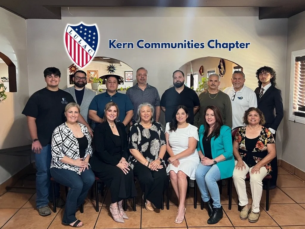 Group photo of people in front of a wall with a logo and text that reads "Kern Communities Chapter." The group includes men and women smiling and sitting or standing in a room with beige walls and wooden flooring.