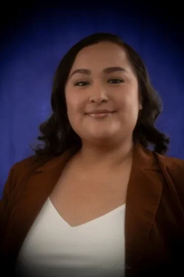 A woman with dark hair smiling in front of a blue background, wearing a brown blazer over a white top.