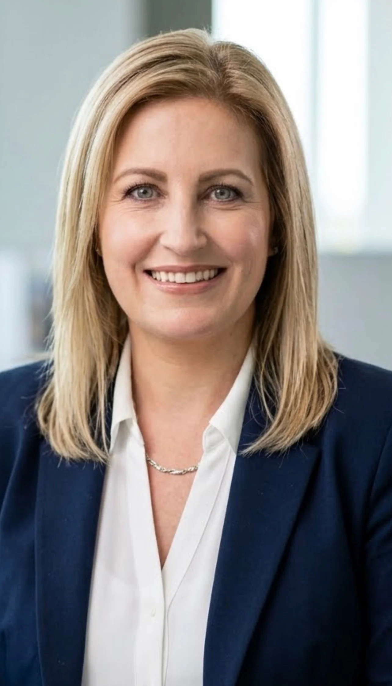 A woman with blonde hair, wearing a white shirt and a navy blazer, smiling confidently in an office setting.