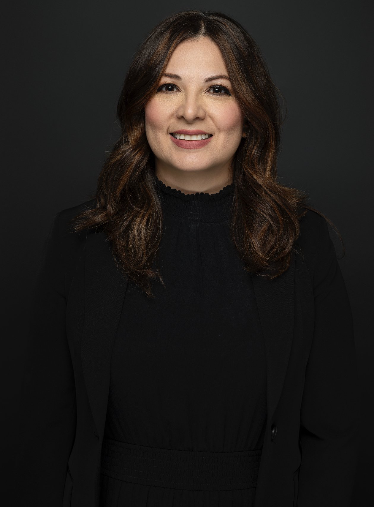Portrait of a smiling woman with shoulder-length brown hair, wearing a black top and blazer, against a dark background.