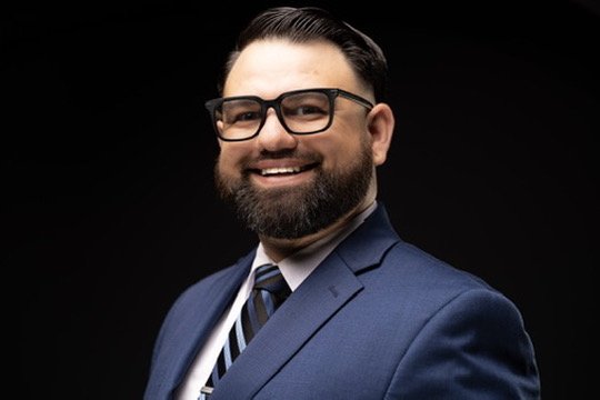A smiling man with a beard and glasses wearing a blue suit and tie, against a black background.