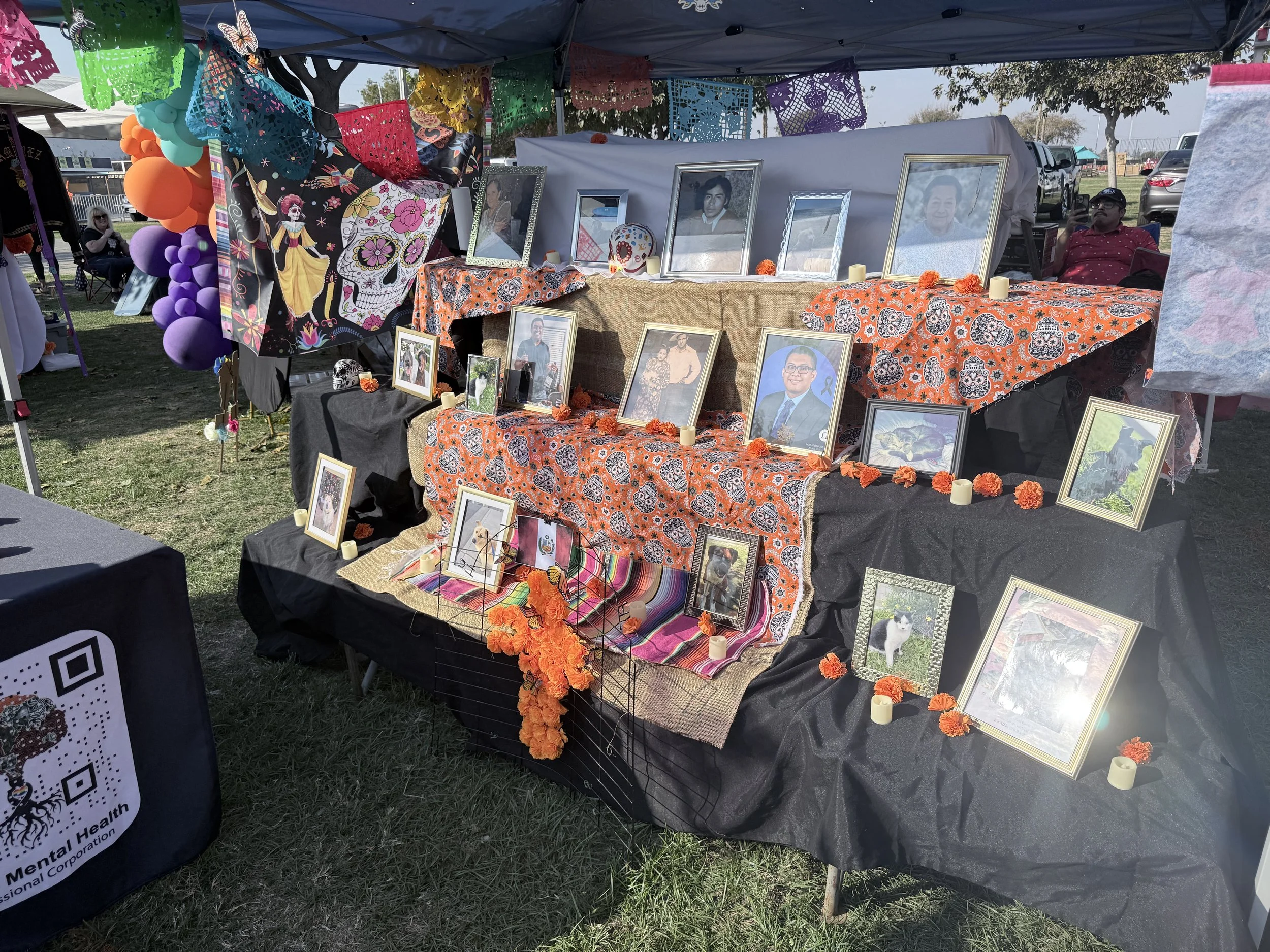 An outdoor altar with framed pictures of loved ones, colorful papel picado banners, orange marigold flowers, and candles, set on tables covered with decorated cloths, part of a Día de los Muertos celebration at a park.