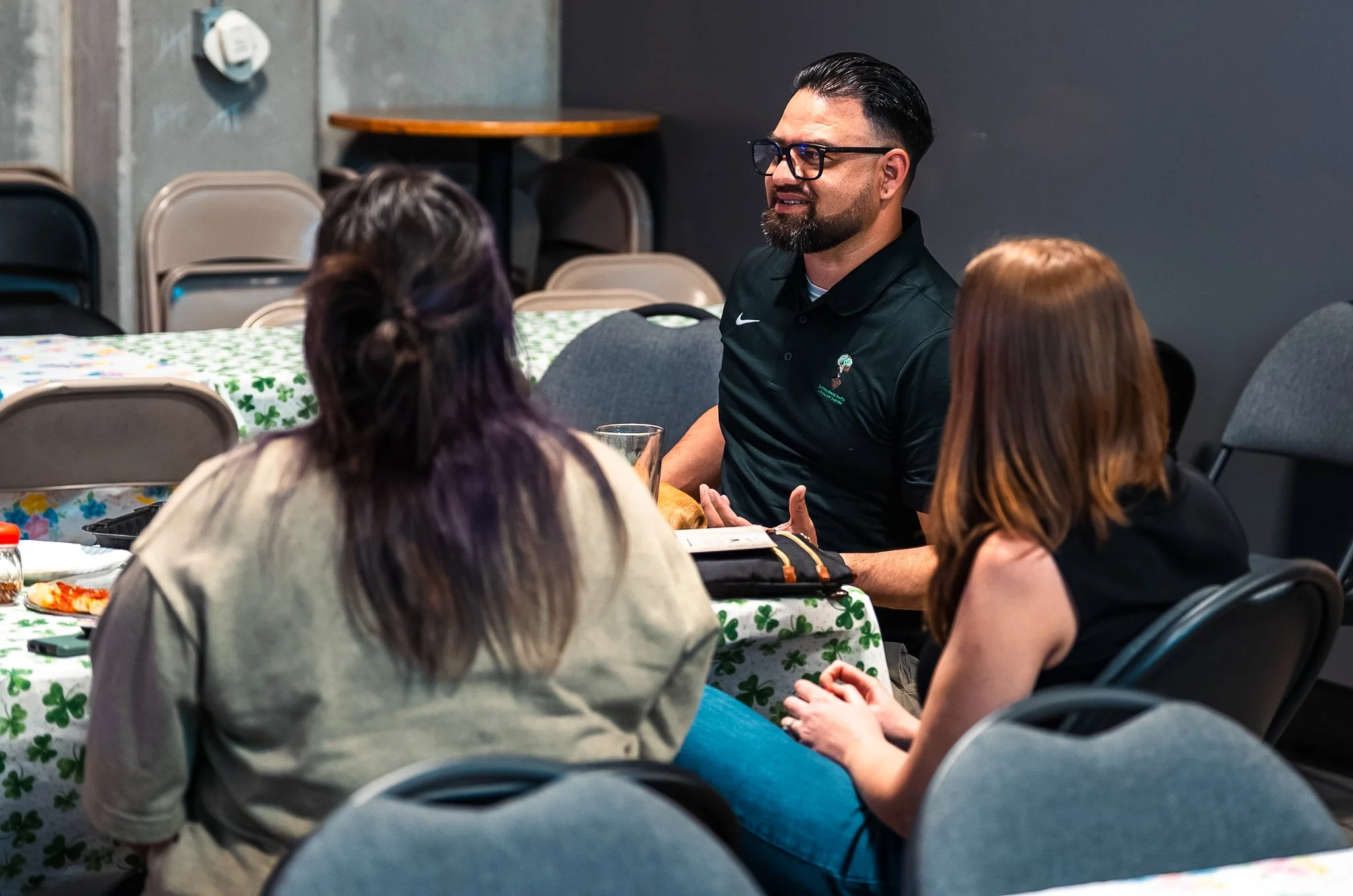 A group of people sitting around a table with a green shamrock tablecloth, engaged in conversation. One man with glasses and a beard is smiling while speaking to the others. The setting appears to be a casual indoor gathering.
