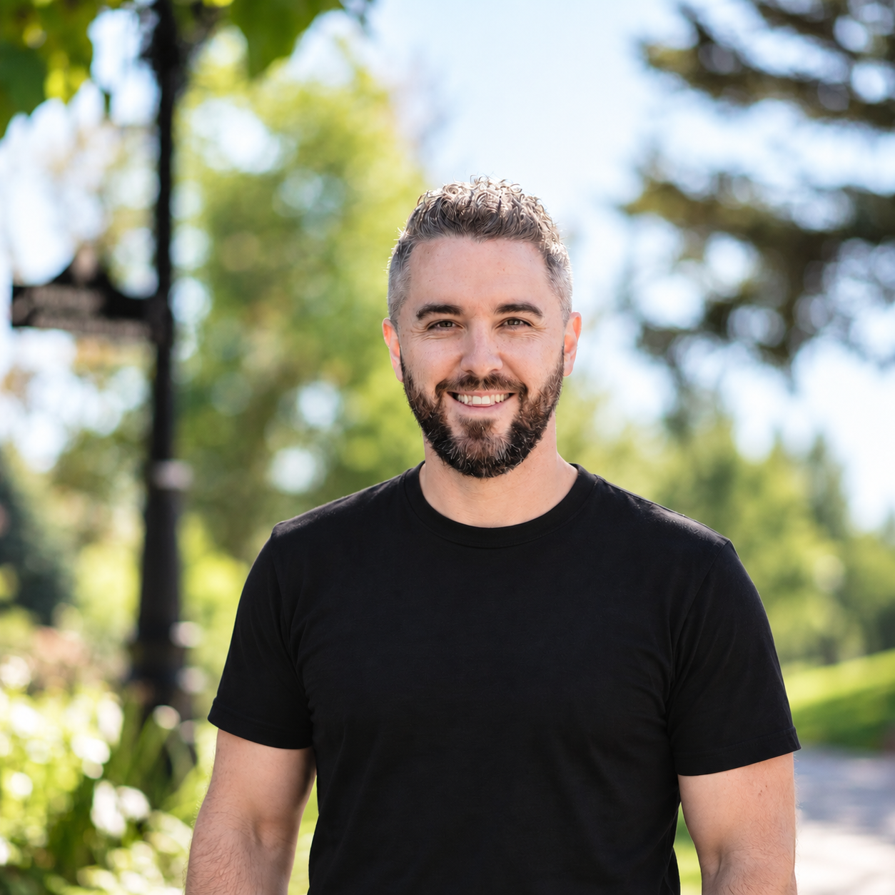 A young man with a beard and short hair smiling outdoors on a sunny day. He is wearing a black t-shirt and standing in a green park with trees in the background.