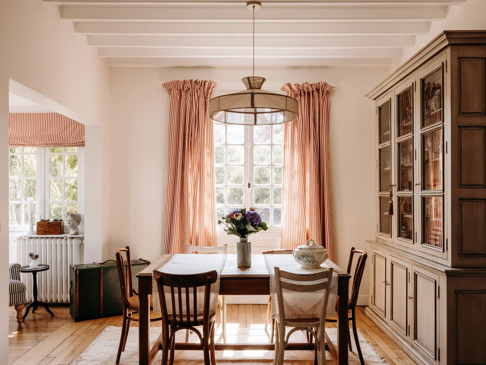 Salle à manger lumineuse avec une grande fenêtre, rideaux à rayures rouges et blanches, une table en bois entourée de chaises anciennes, une plante avec des fleurs au centre, et un buffet en bois sur le côté droit.