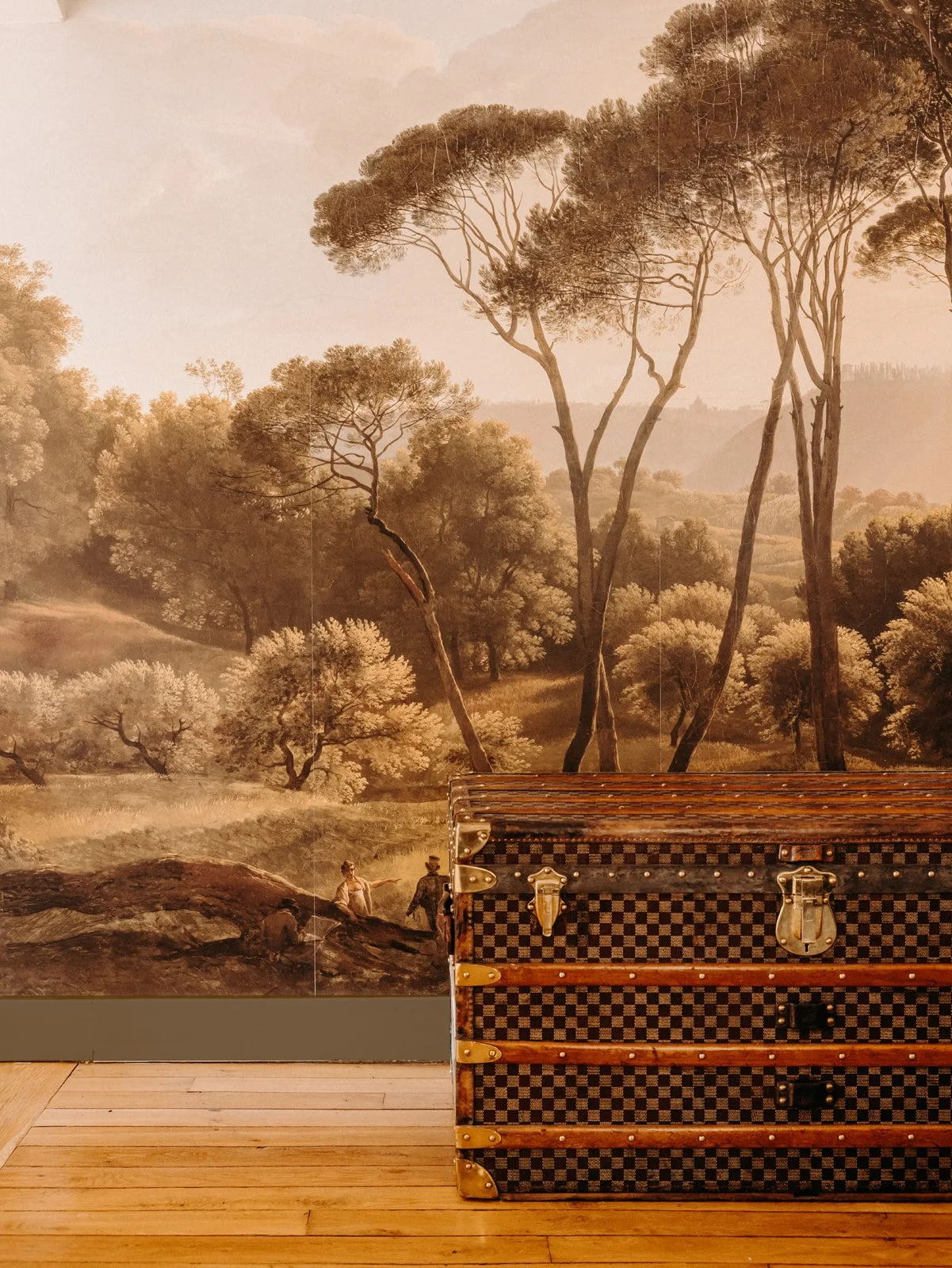 Peinture murale représentant une forêt avec des arbres et des collines. En bas, deux personnes discutent près d'une roche, à côté d'une valise en bois.