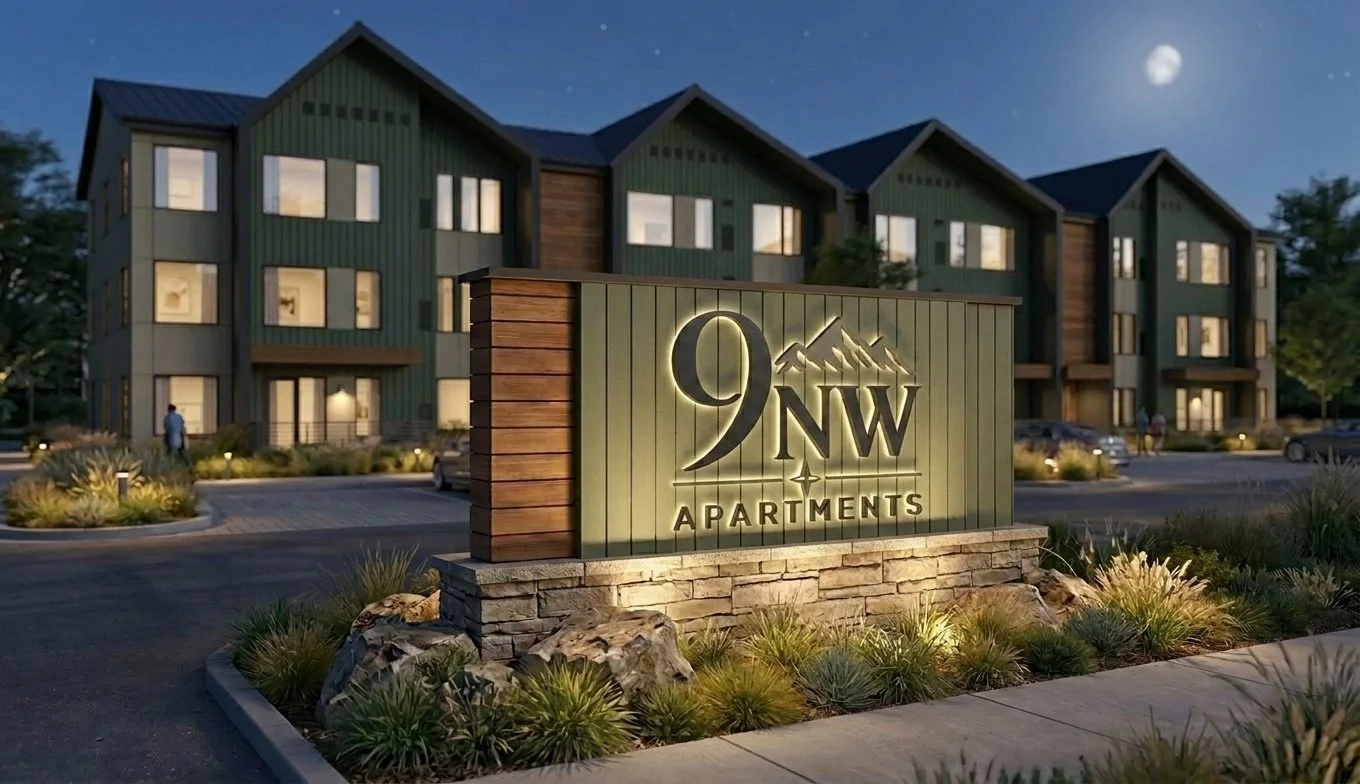 Night view of a modern apartment complex with a illuminated sign reading "9 NW Apartments" in the foreground, landscaping with rocks and plants, and a dark sky with the moon.