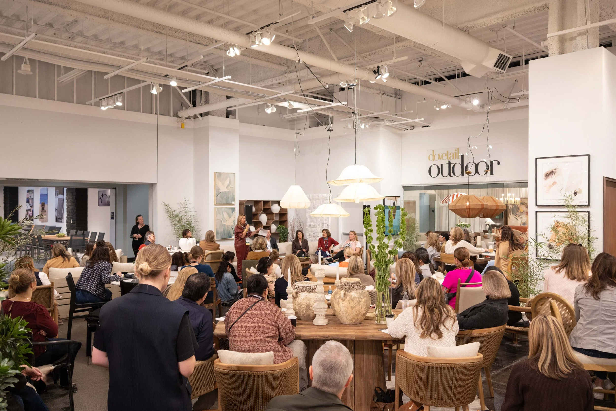 A large group of people seated in a spacious interior with modern decor, attending a presentation or seminar. There is a woman speaking at the front with a microphone, surrounded by framed artwork, potted plants, and hanging lamps.