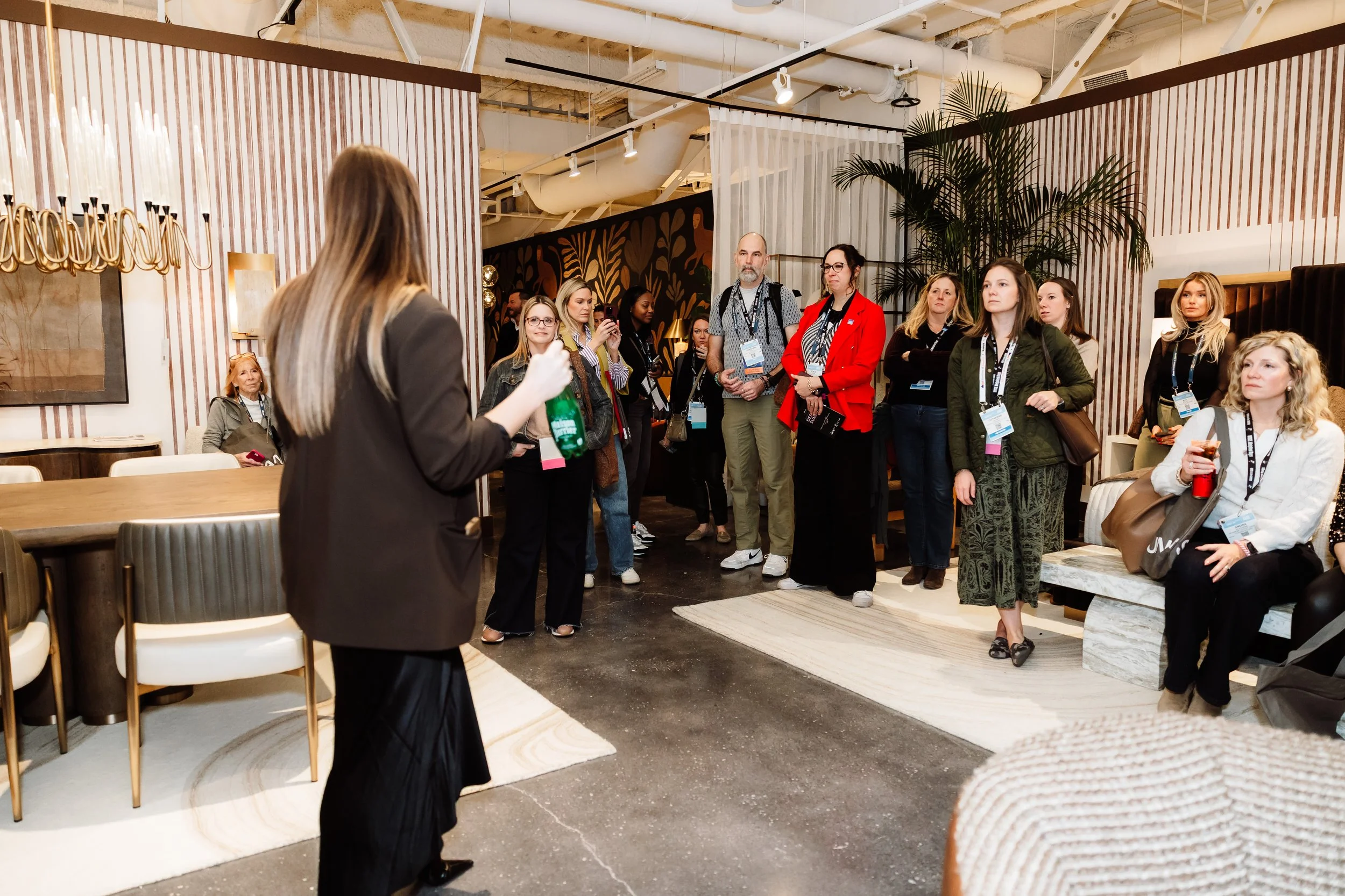 A group of diverse people standing and sitting in a modern, well-lit indoor space, listening to a speaker in the foreground. The audience includes men and women of different ages, some holding drinks and others taking photos or looking attentively.