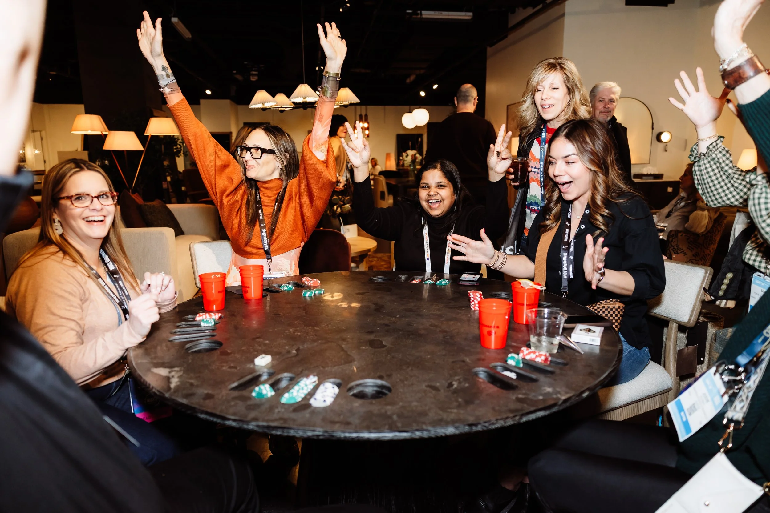 A group of women playing poker at a round table, celebrating, smiling, with poker chips and cards on the table in a cozy, well-lit room.