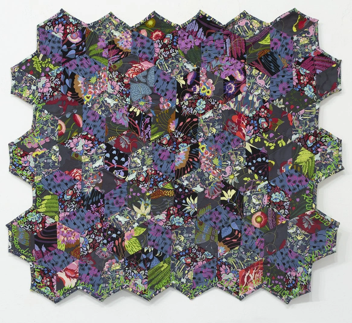 Small quilt with hexagonal design in gray, blue, and purple floral fabric, with green text around edges reading, "I wish I could tell you it's as simple as spending less than you earn, but that only works if you're able to earn enough to begin with,