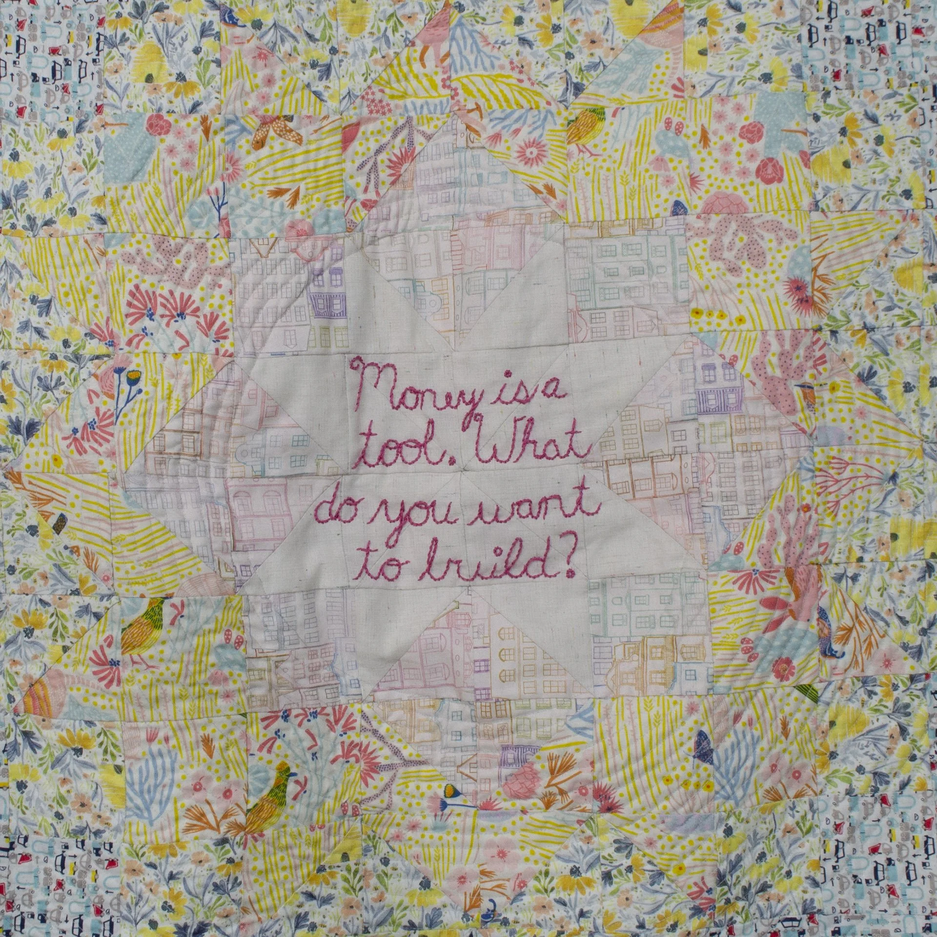 Detail of quilt with warm tones and the embroidered words, "Money is a tool. What do you want to build?"