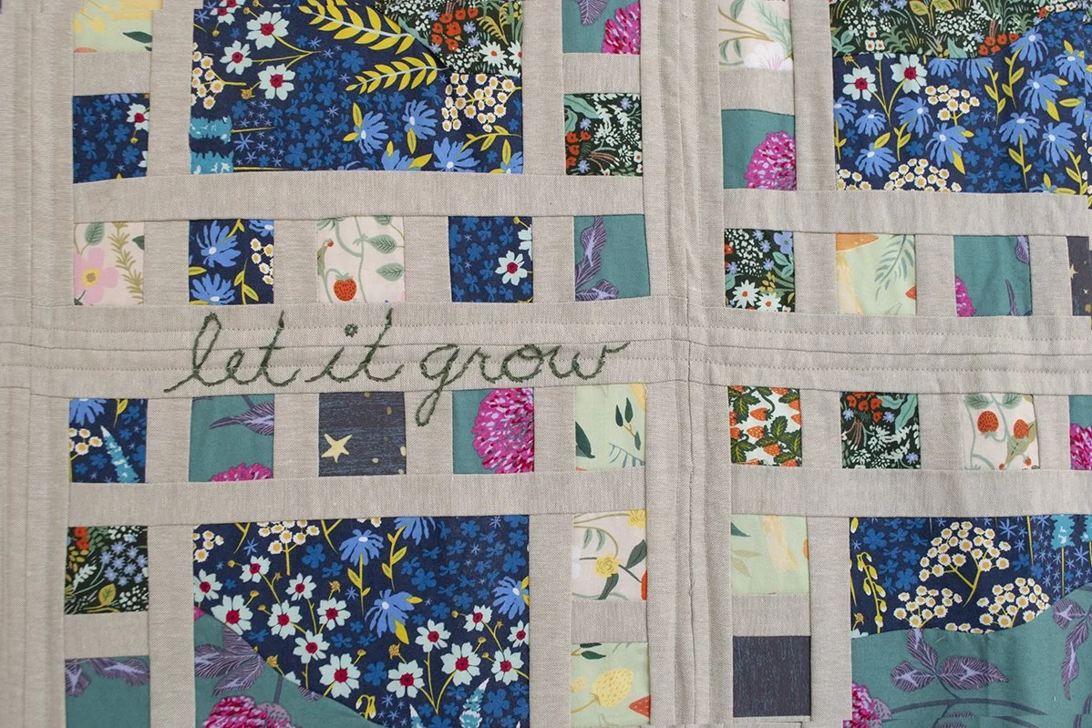 Detail of quilt with linen background and many colorful squares with floral designs and the embroidered words "let it grow"