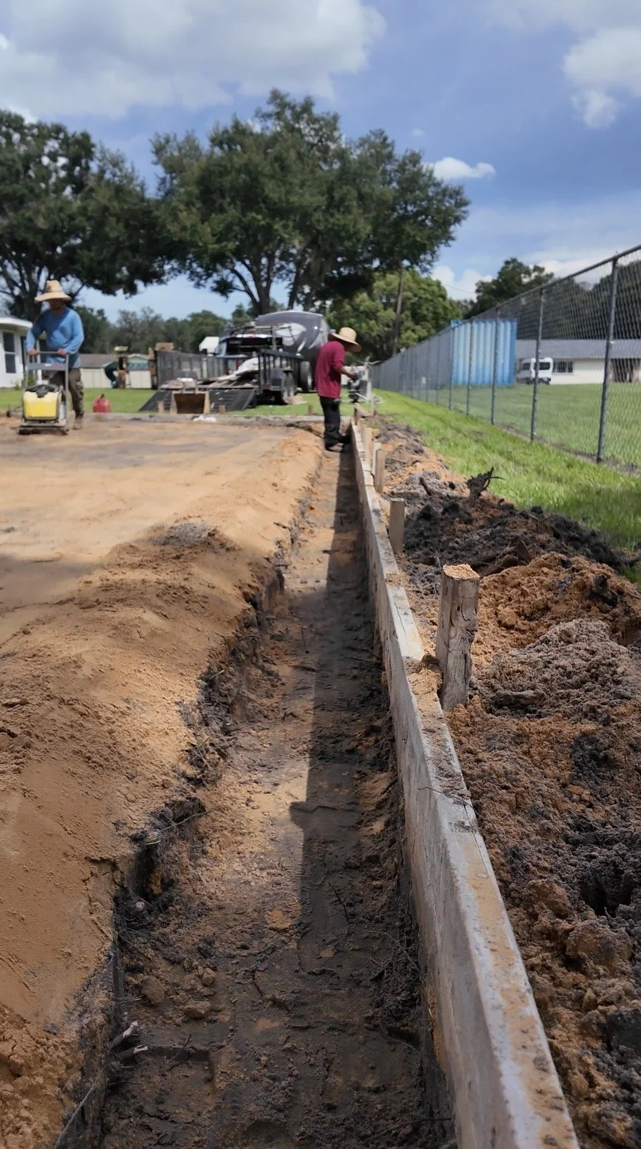 Workers constructing wooden form and trench, on a sunny day with trees and a chain-link fence in the background.