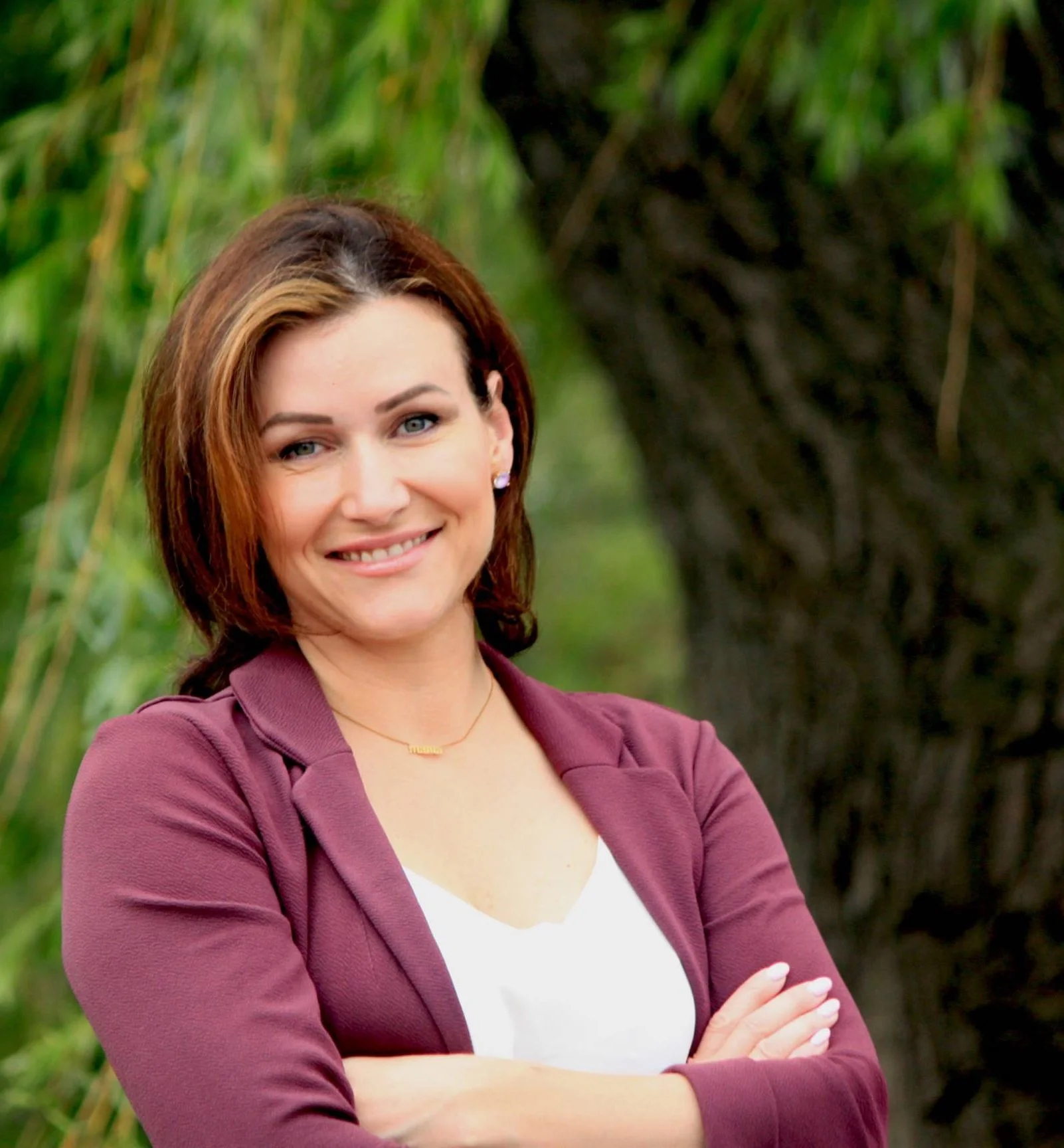 A woman with short auburn hair and blue eyes smiling outdoors, wearing a purple blazer over a white top, standing in front of a background of green trees.