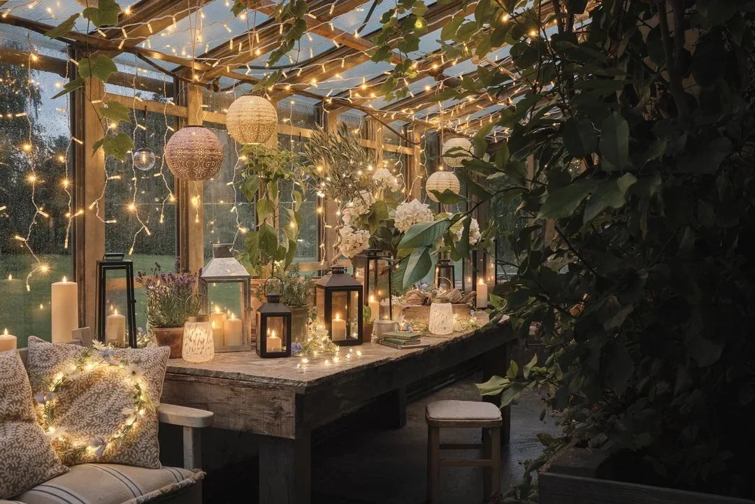A cozy greenhouse decorated for evening with string lights, candles, lanterns, and hanging paper lanterns. There are plants and flowers on the table and around the space, with a cushioned seating area with pillows on the left.