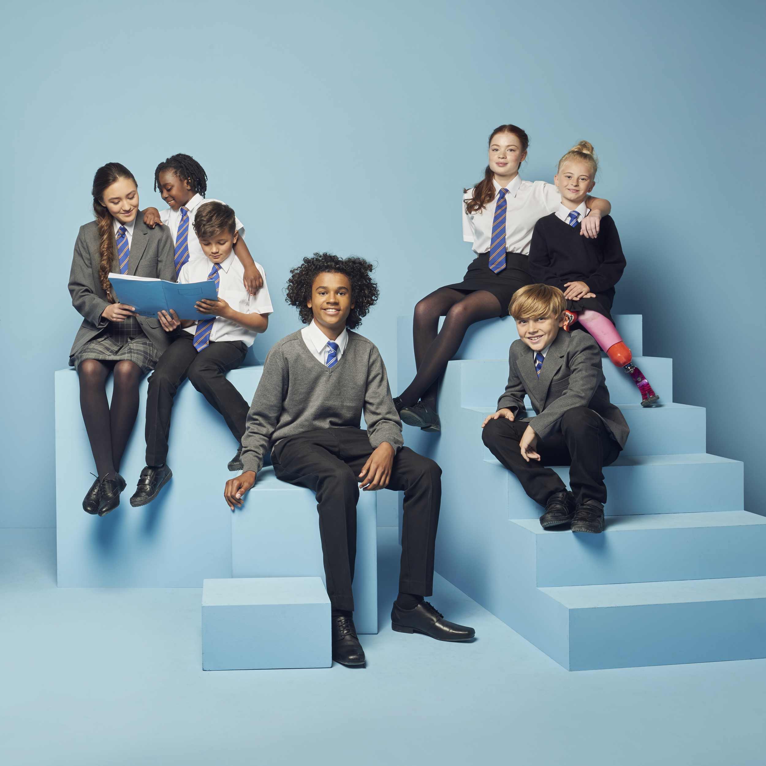 Group of primary & Secondary school children in uniforms with red, pink, and purple leggings, sitting and standing on blue steps against a blue background, some looking at a book and others smiling.