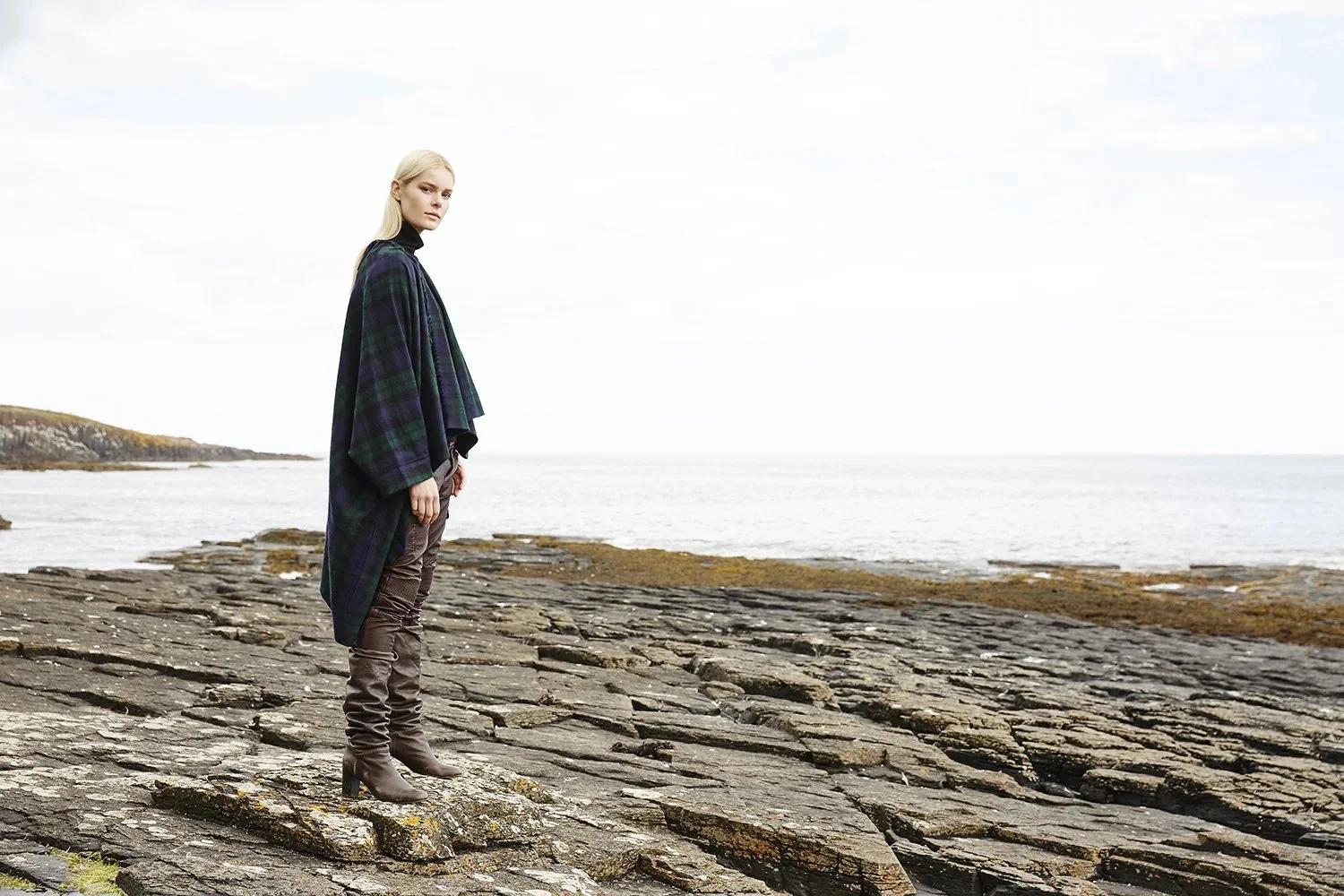 A woman with long blonde hair standing on a rocky shoreline near the ocean, wearing a dark plaid cape and brown heeled boots, looking at the camera with a neutral expression.