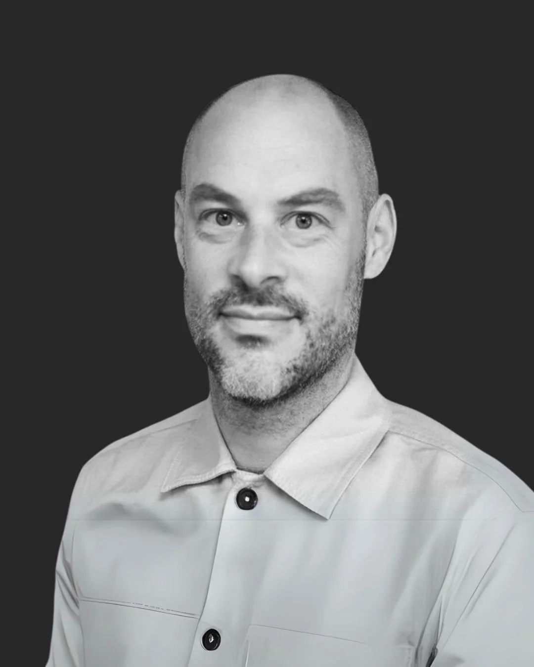 Black and white professional portrait of a man with a bald head, beard, and light-colored collared shirt, set against a dark background.
