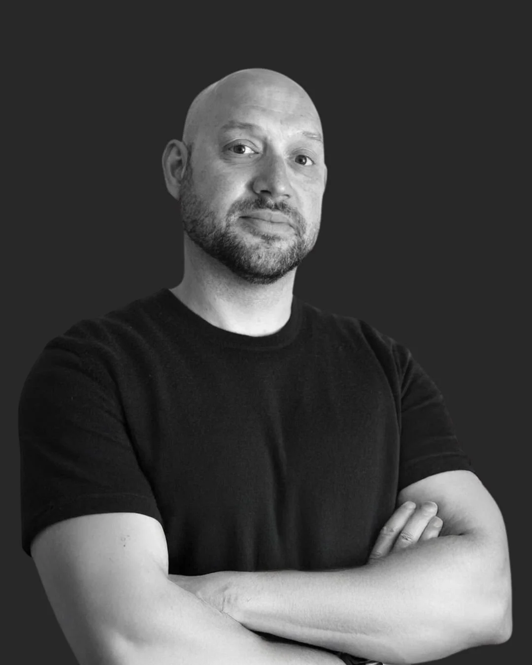 Black and white portrait of a man with a bald head and beard, wearing a black t-shirt, standing with arms crossed against a dark background.
