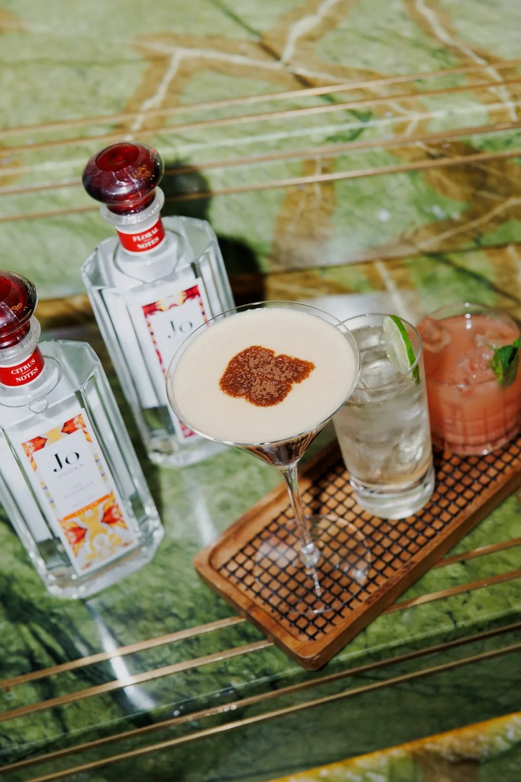 A cocktail glass with a cream-colored beverage garnished with a sprinkle of cinnamon, a bottle of Jo Notes citrus-flavored spirit, a glass with ice and lime wedge, and another glass with a pink drink and mint, all on a wooden tray on a green and brown textured surface.