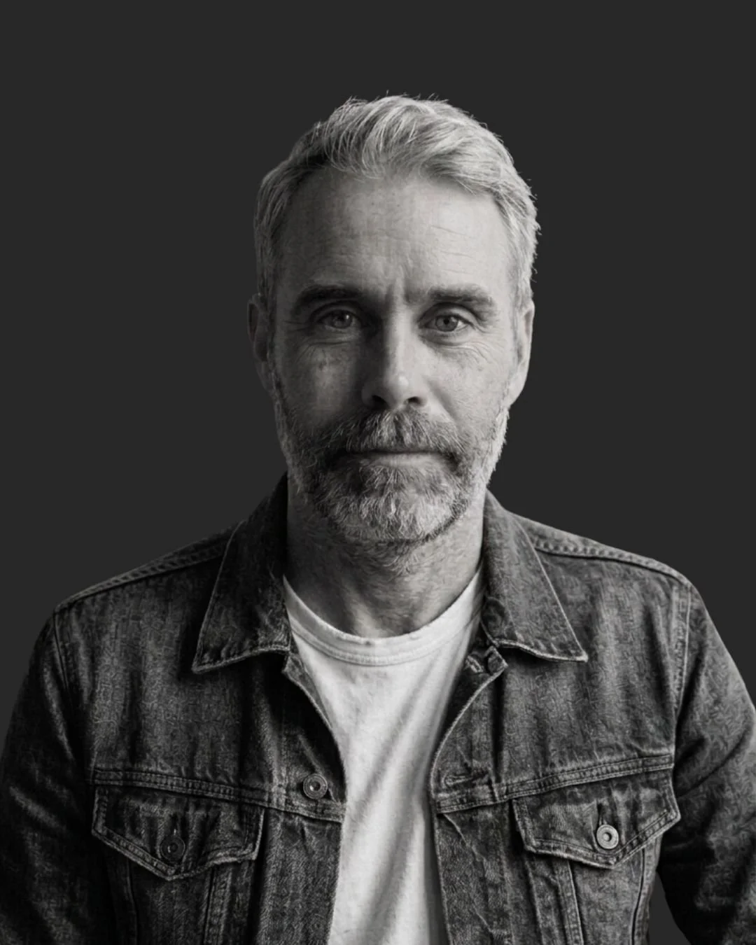 Black and white portrait of a middle-aged man with gray hair, beard, and mustache, wearing a denim jacket over a light-colored shirt, against a dark background.