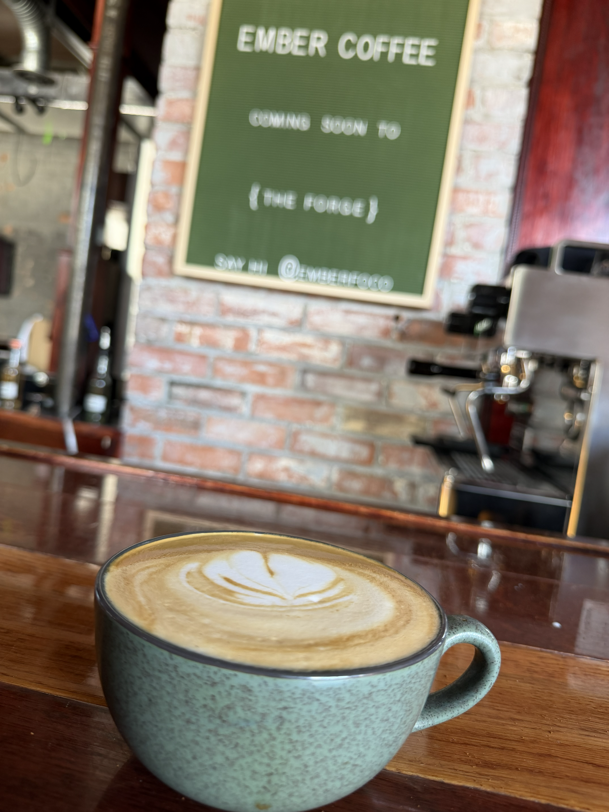A cup of coffee with latte art on a wooden table in a coffee shop with brick walls and a green sign in the background.