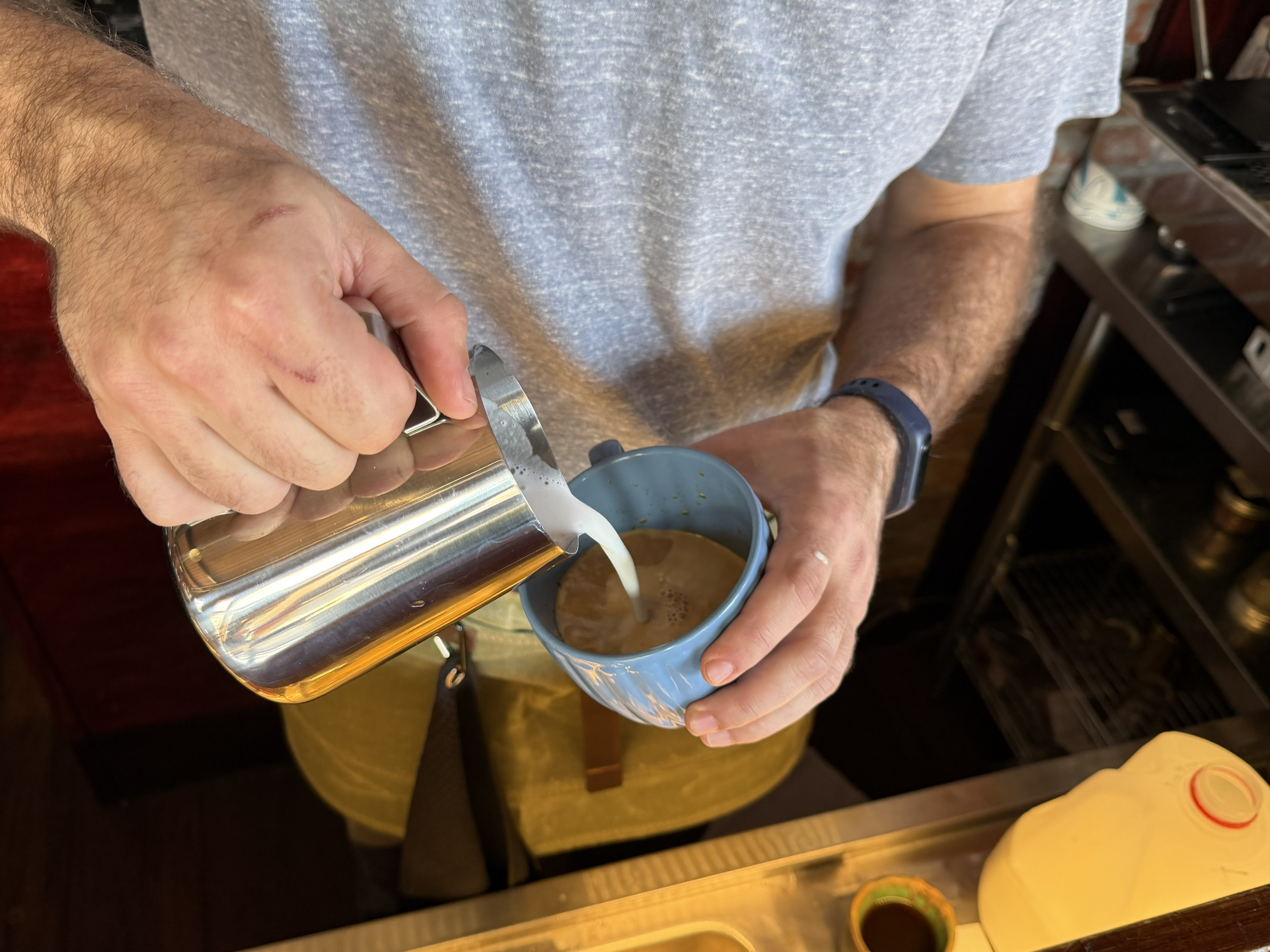 A person wearing a gray T-shirt and a black watch is pouring a light-colored liquid, possibly coffee, from a silver carafe into a small blue mug.